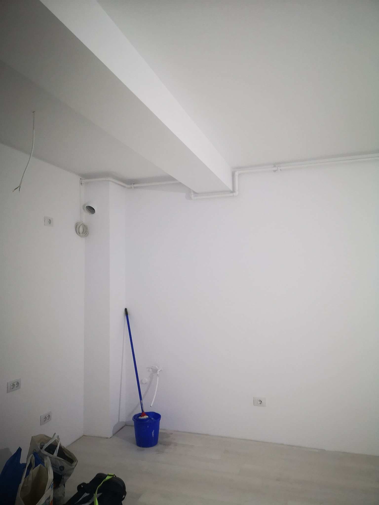 Empty room with white walls and ceiling, several electrical outlets, a blue bucket with mop, some bags and tools on the floor, and visible PVC piping along the ceiling.