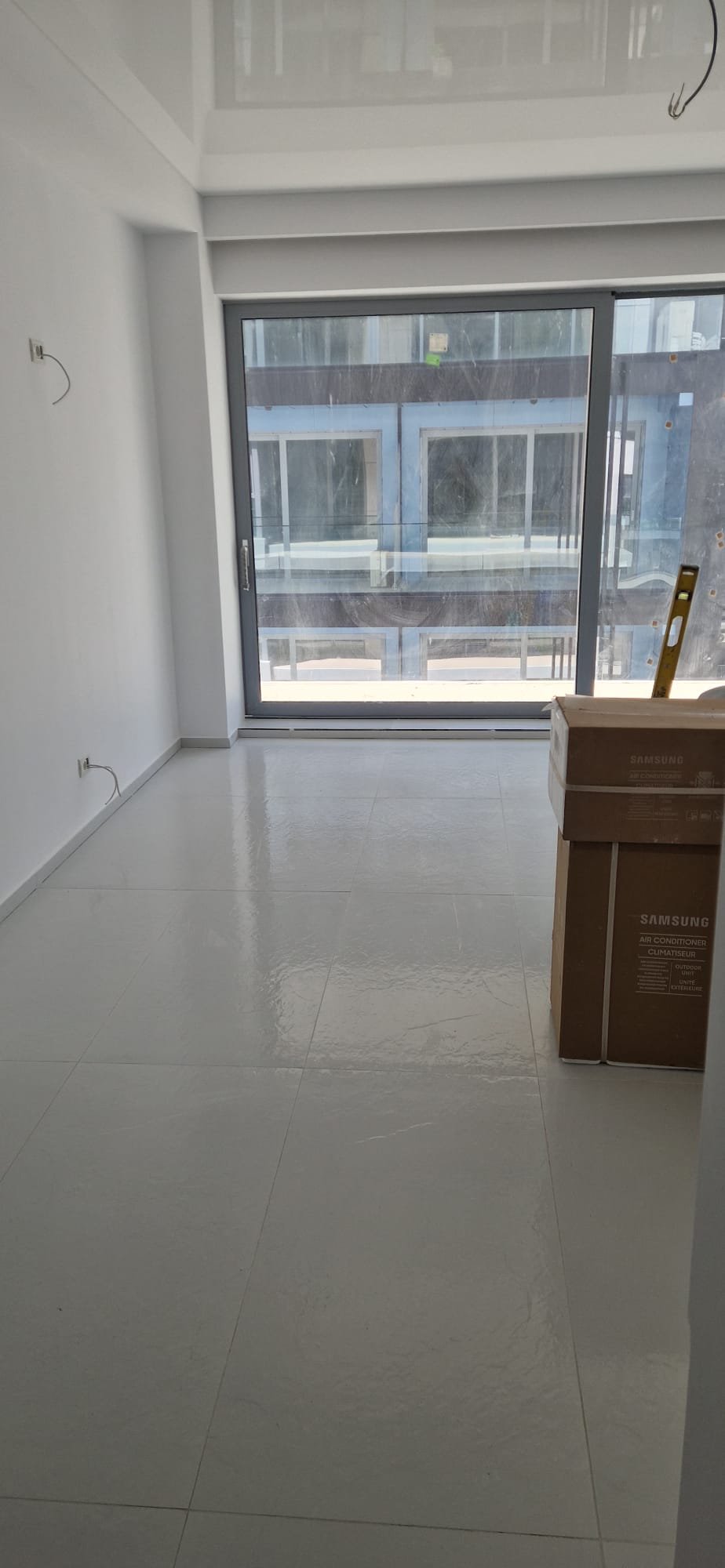 An empty room with white tiled floors, white walls, and a large sliding glass door leading to a balcony. There are construction boxes and a ladder inside the room.