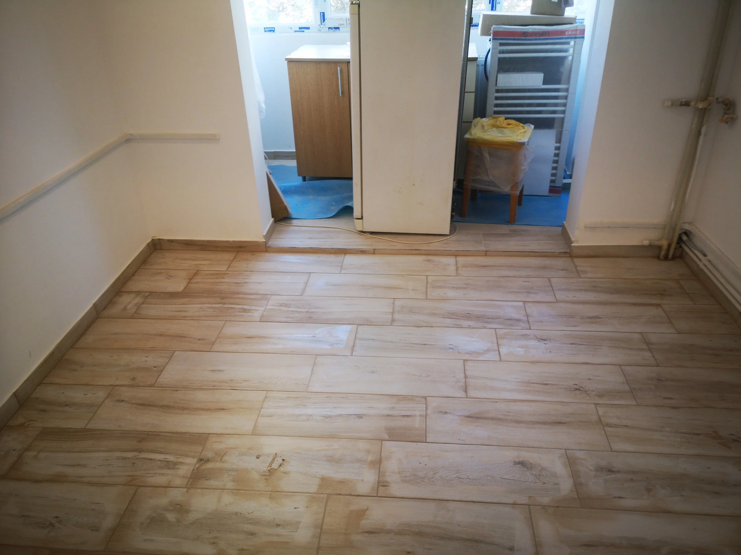View of a room with wooden flooring, white walls, and a doorway leading to a utility or laundry area containing a fridge, a small table with a yellow cloth, a rack, and various boxes and items.