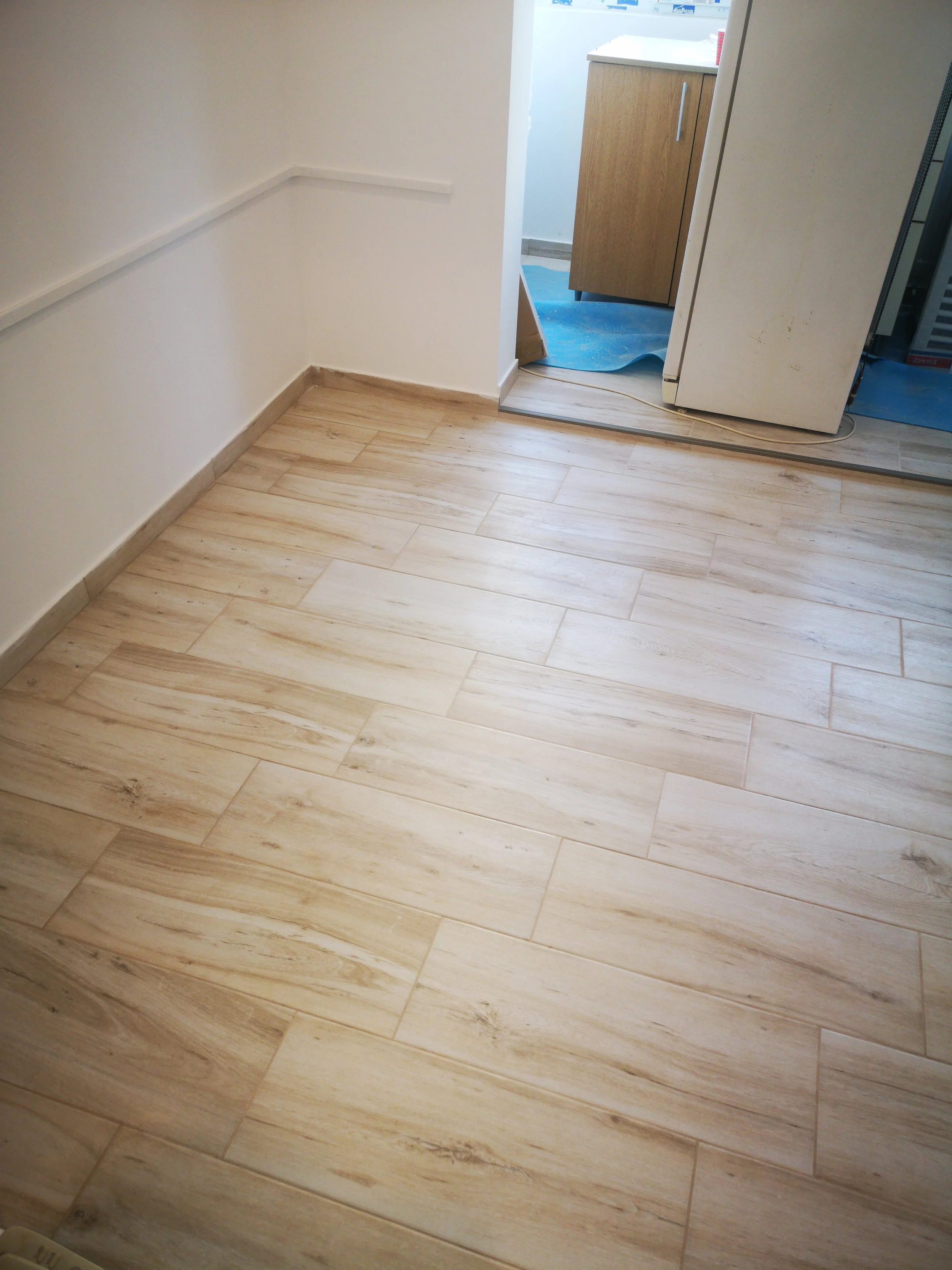 An interior space with light-colored wood-look tile flooring, white walls, and a partial view of a small kitchen area with a wooden cabinet, a refrigerator, and some blue protective floor covering.