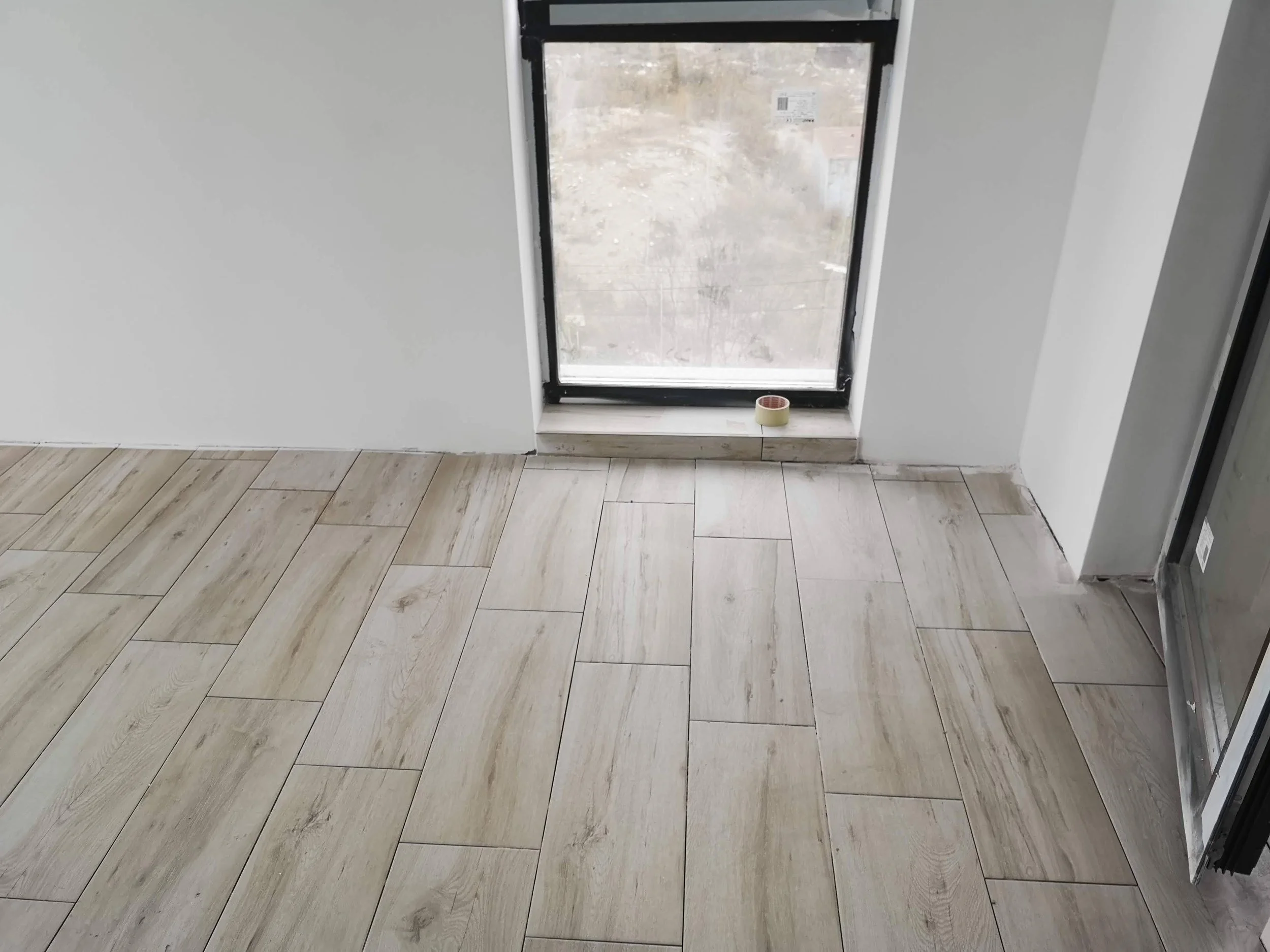Interior of a room with a large window, light wood tile flooring, white walls, and a roll of masking tape on the windowsill.