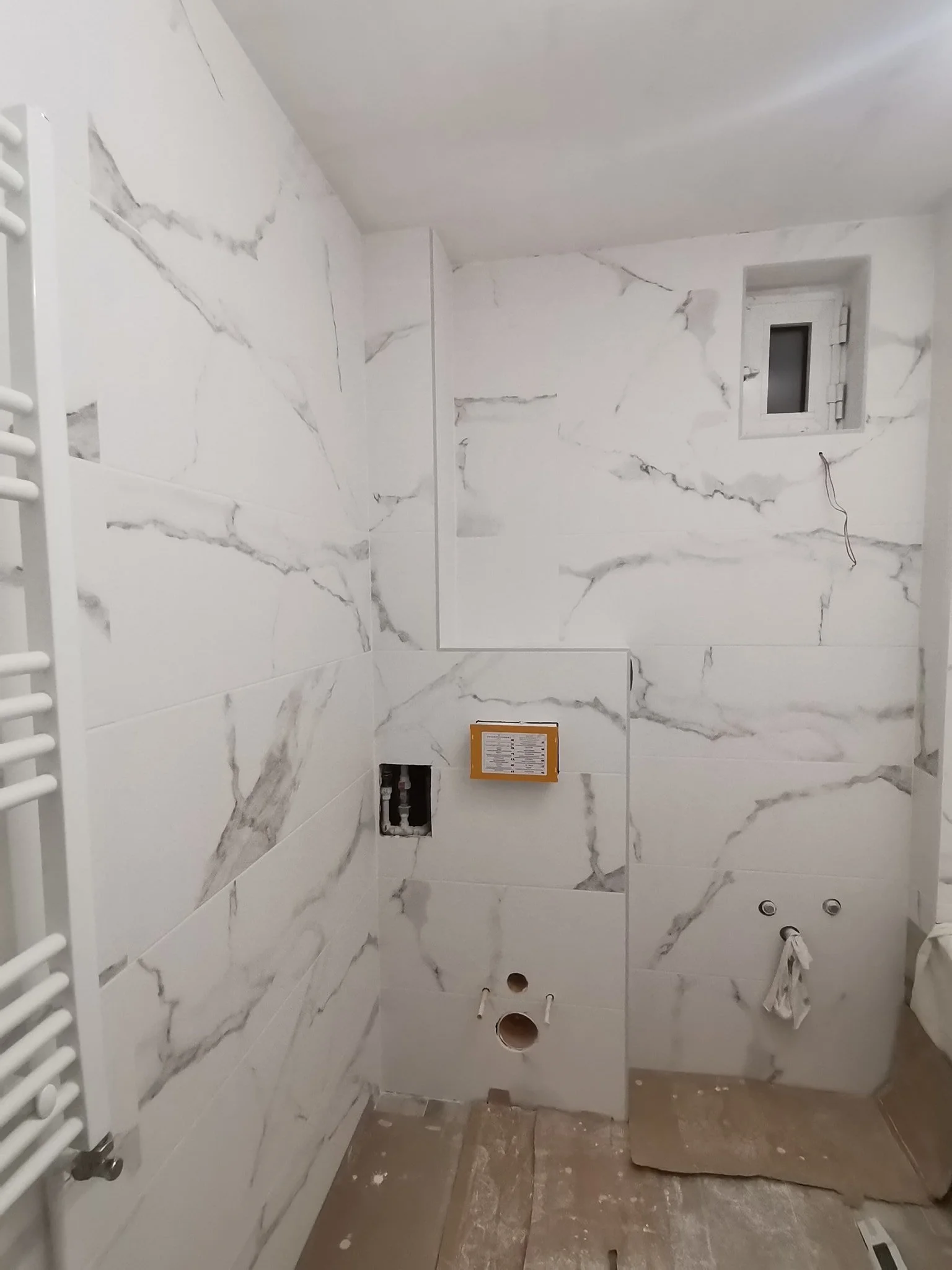 A bathroom under construction with white marble-like tiled walls, a small window, and exposed plumbing openings.