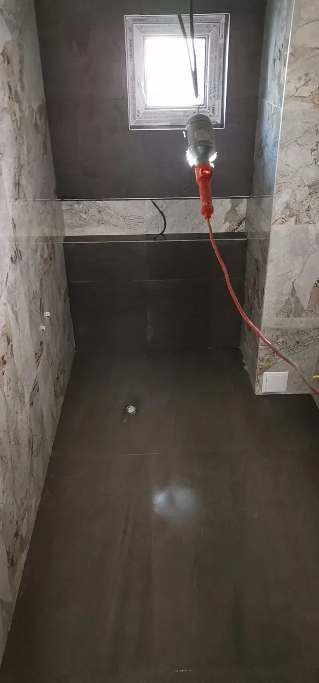 Bathroom under construction with a small window, a red electrical cord hanging from the ceiling, and a corner with tiled walls and floor.