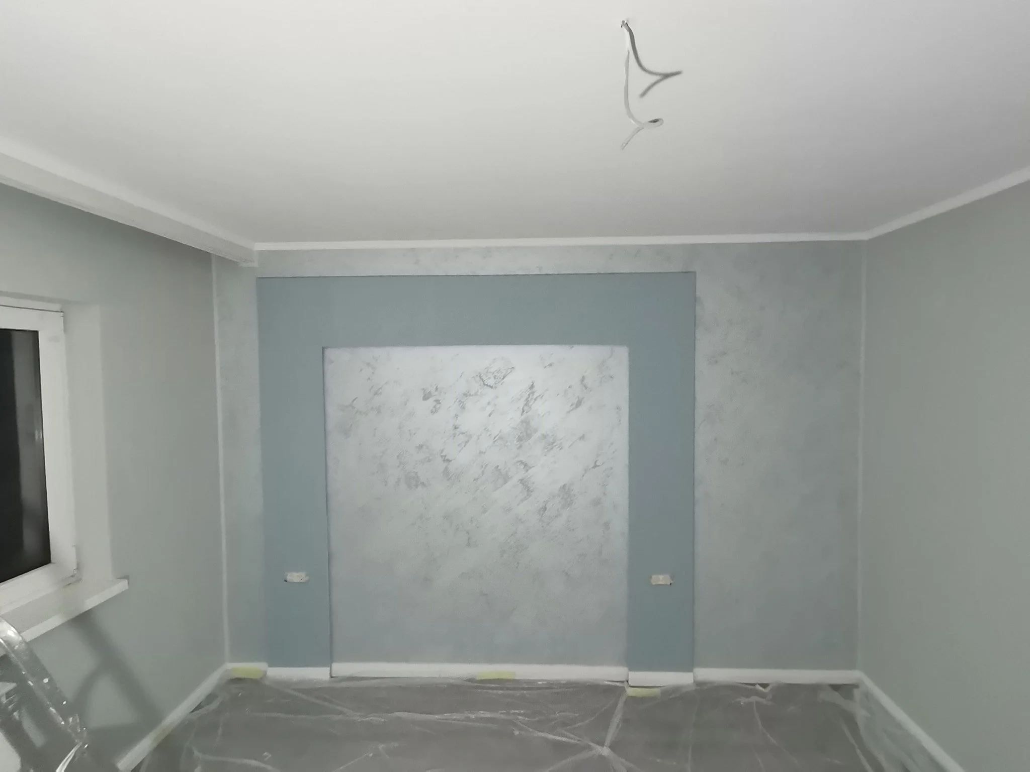 A room under renovation with gray walls, a white window frame, and a ceiling with exposed wires hanging down. The floor is covered with plastic protective sheeting.