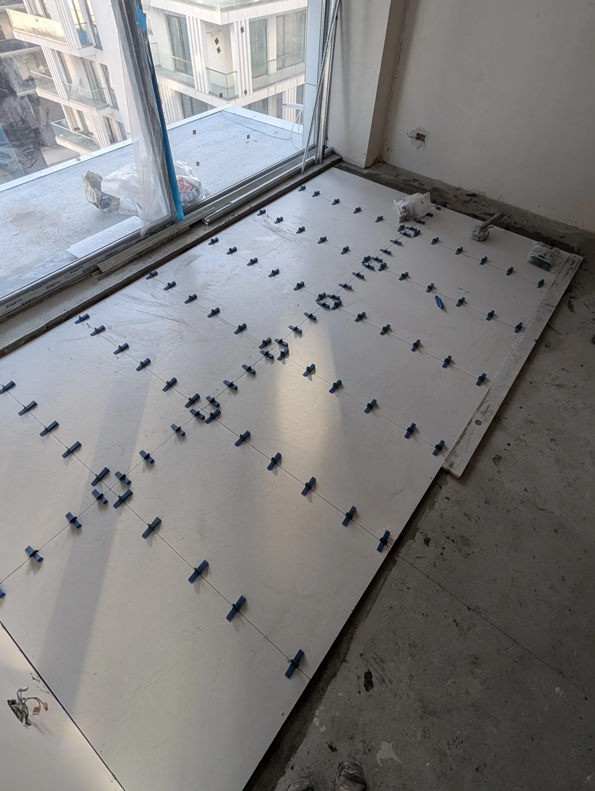 Interior of a room under construction with white floor tiles being installed, held in place with spacers, near a large glass sliding door leading to a balcony.