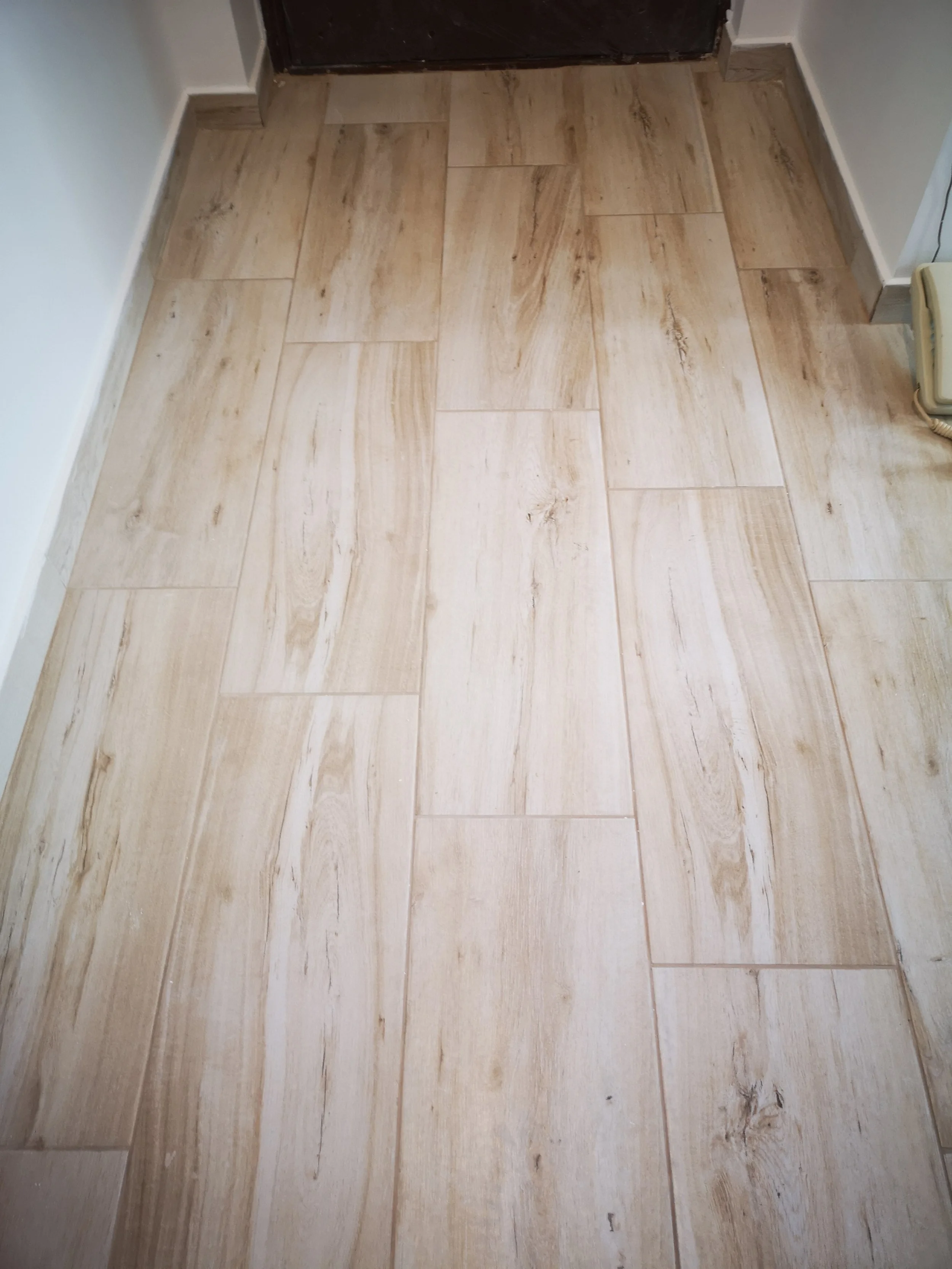 A wooden floor area in front of a dark-colored door, with a white wall on either side and a baseboard trim.
