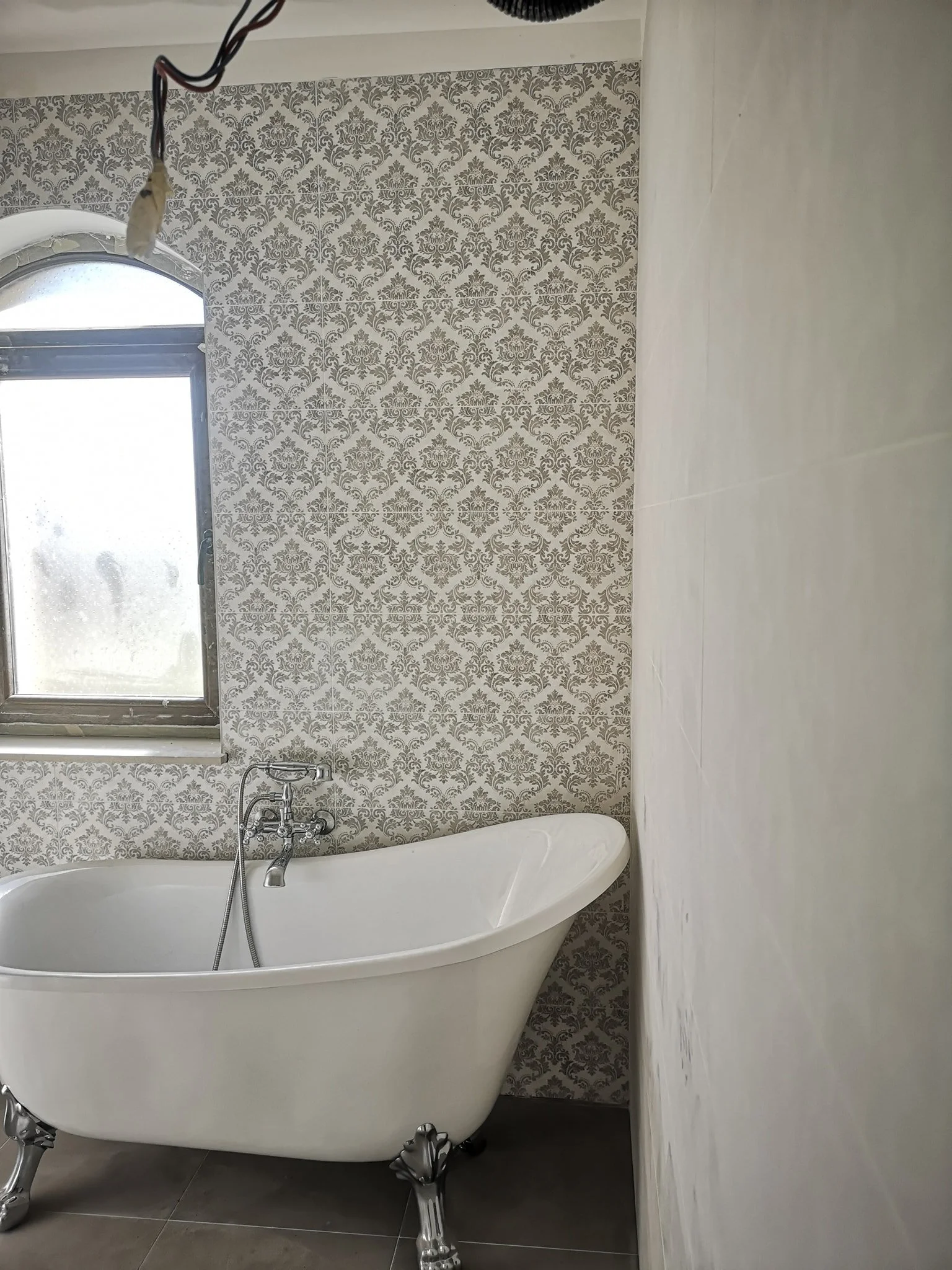 A vintage white clawfoot bathtub with silver clawfoot legs in a bathroom, with patterned wallpaper and a window.