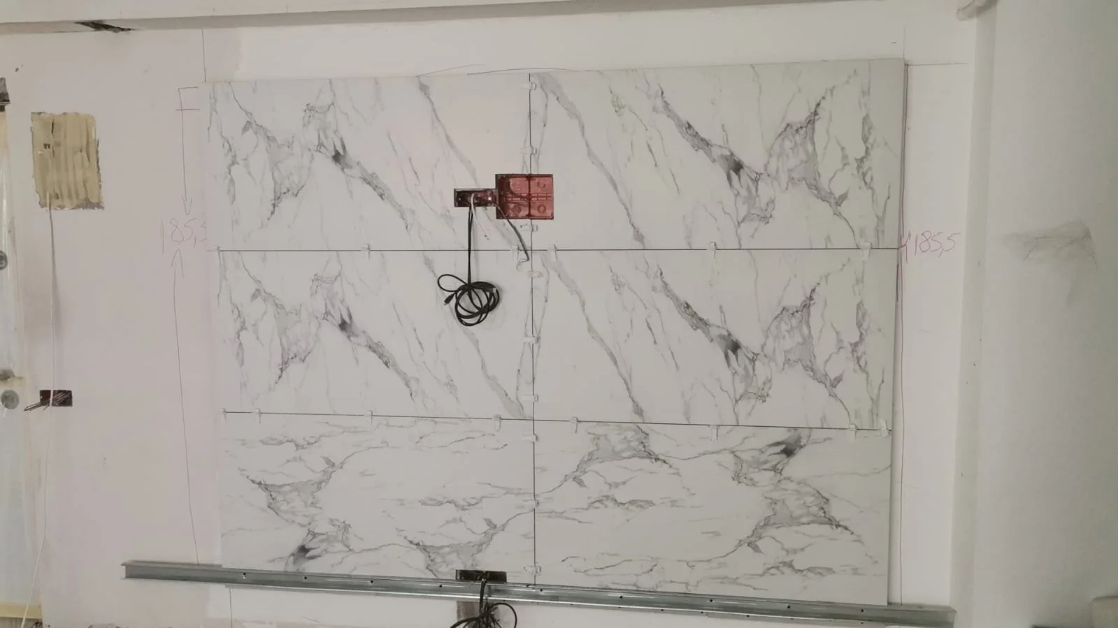 Large white marble tile with gray veining installation in progress on a wall; marked measurements in pink, wiring and electrical components present.