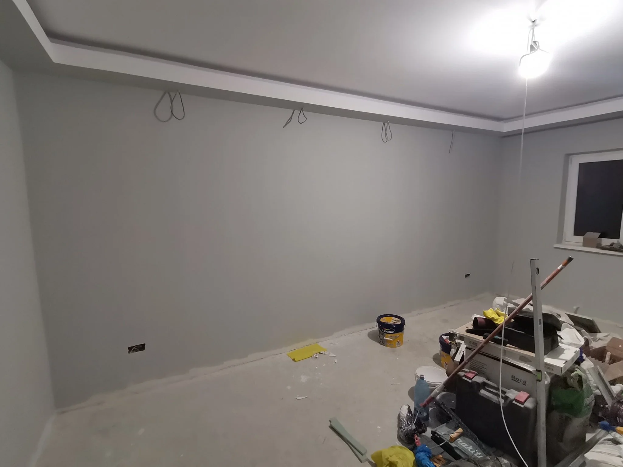 A room under construction or renovation with unpainted light gray walls and various tools and materials scattered on the floor, including a yellow bucket, paint cans, and construction supplies, with electrical wiring hanging from the ceiling.