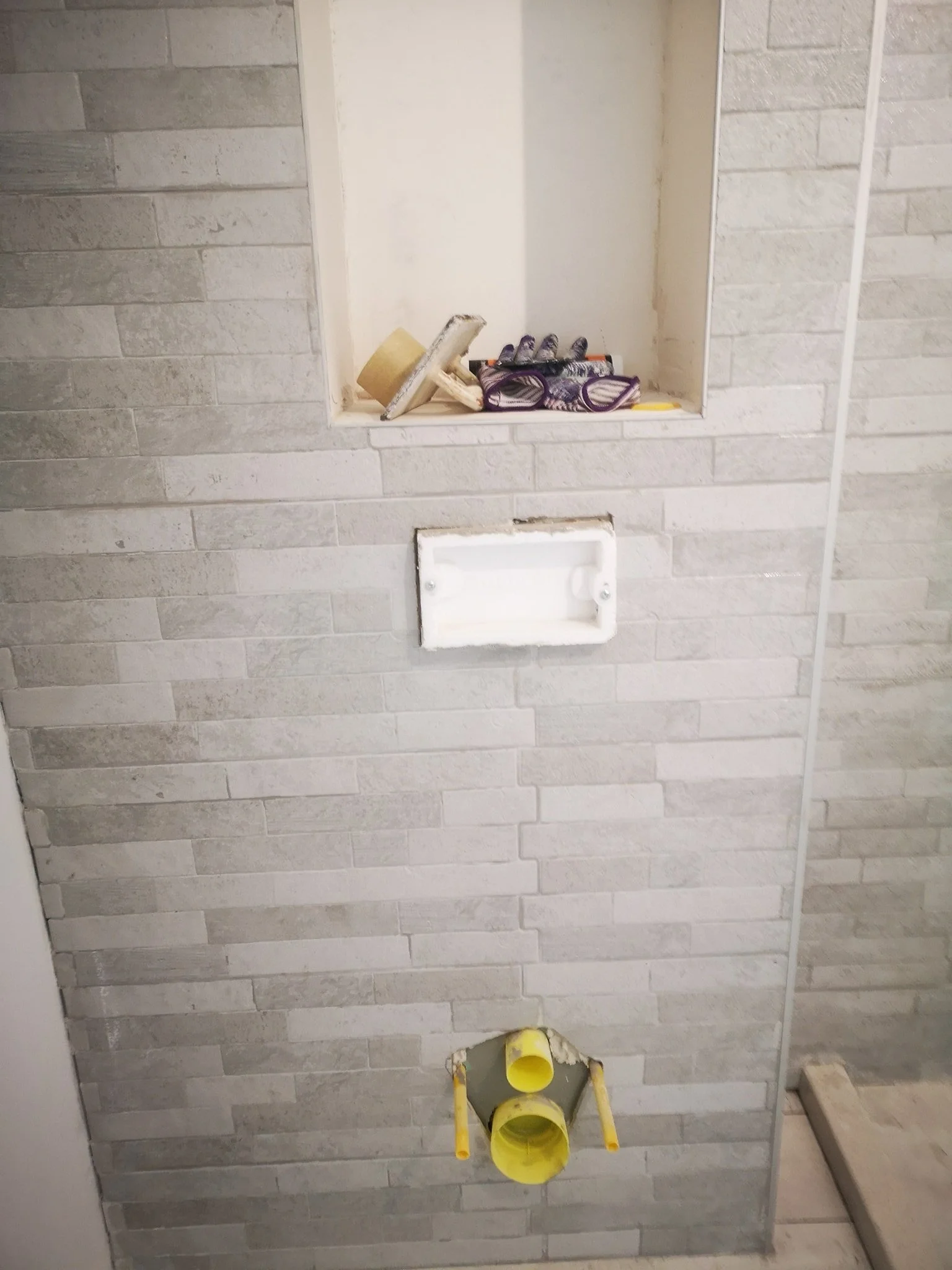 Bathroom wall with unfinished plumbing and construction work, including a yellow PVC pipe, a hole in the wall, and a construction panel on the floor.