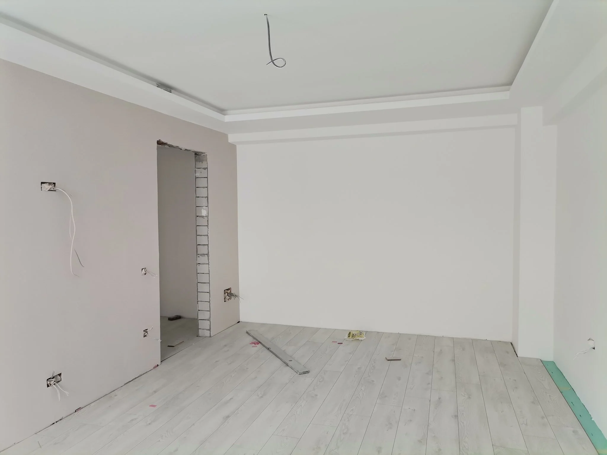 Interior room under construction with white walls, partially installed flooring, and an unfinished doorway with exposed bricks. Wiring is visible on the walls and ceiling.