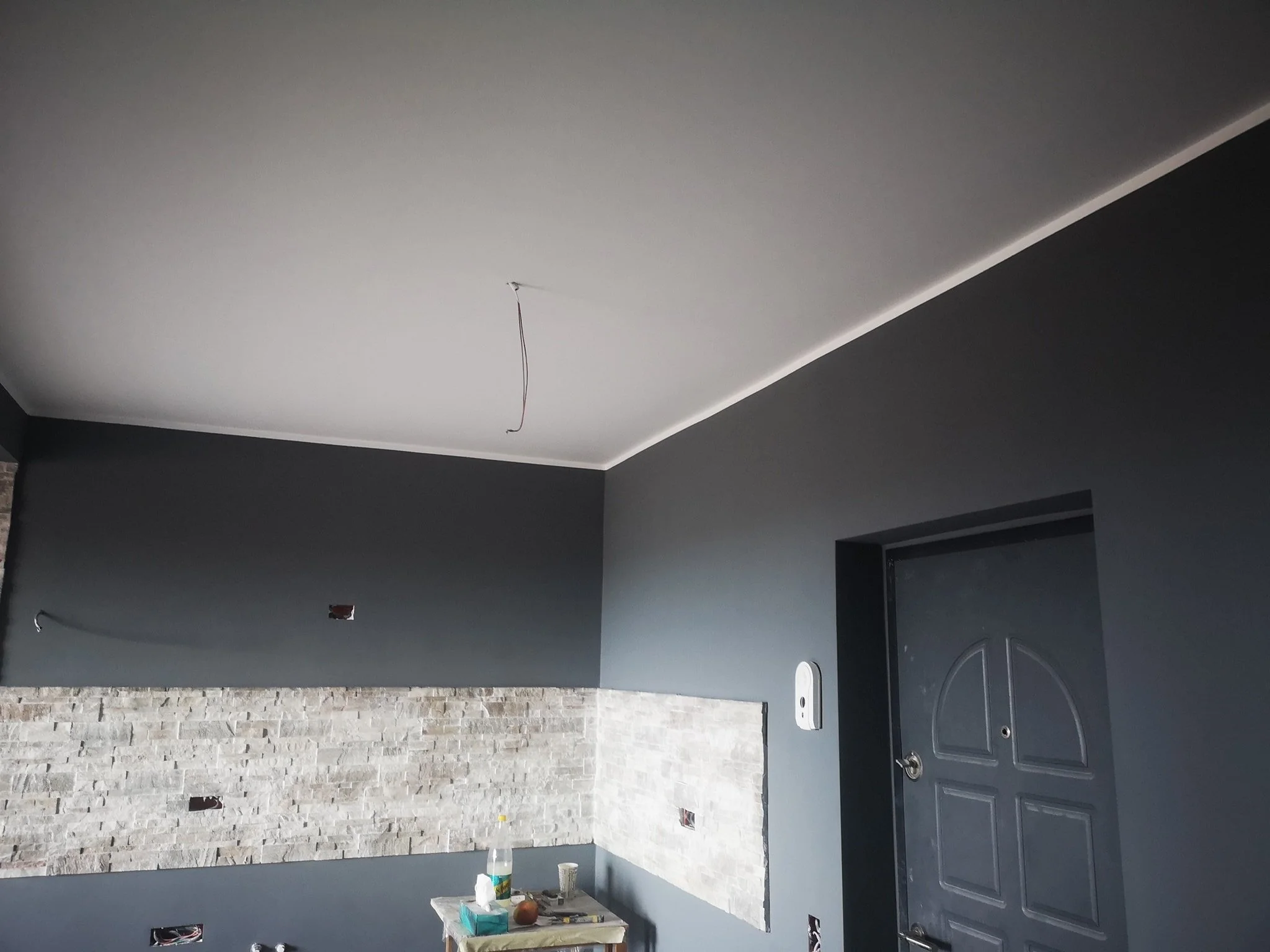 Interior of a room under construction or renovation, showing a ceiling with a wire hanging down, a textured stone wall, and a partially fitted sink with a faucet.