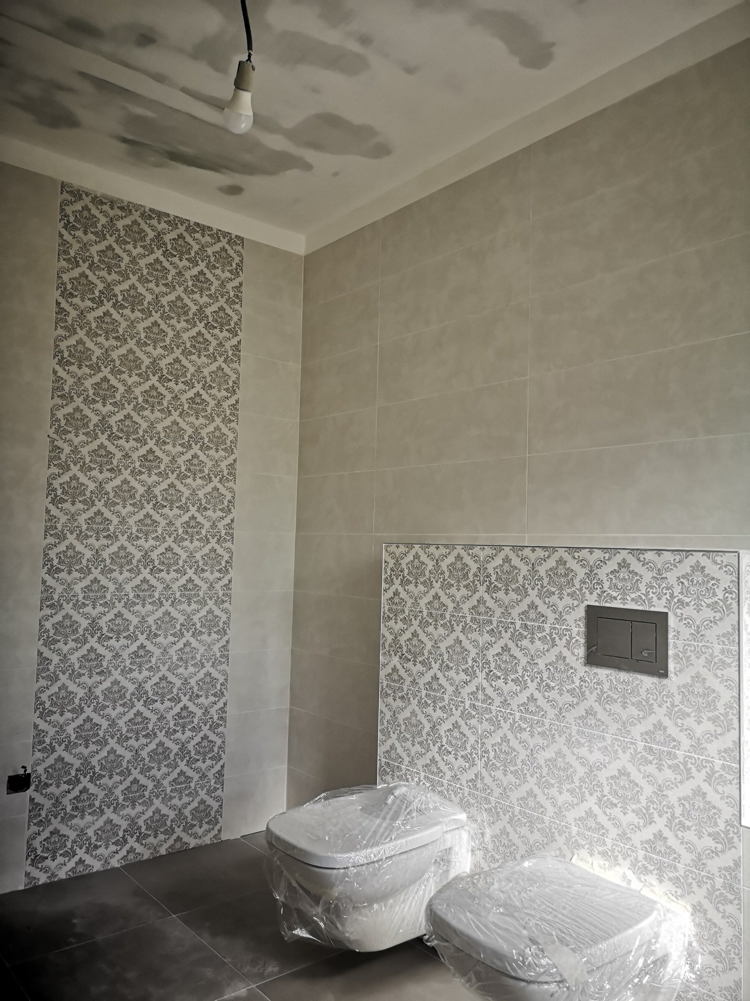 A bathroom under construction with a toilet covered in plastic, tiled walls with decorative patterns, and an exposed light bulb hanging from the ceiling.