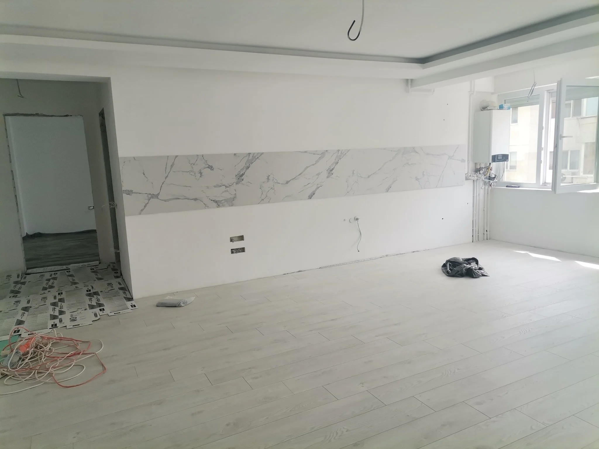 An unfinished room with light wooden flooring, partially painted white walls, a large window, and some exposed wires and construction materials on the floor.
