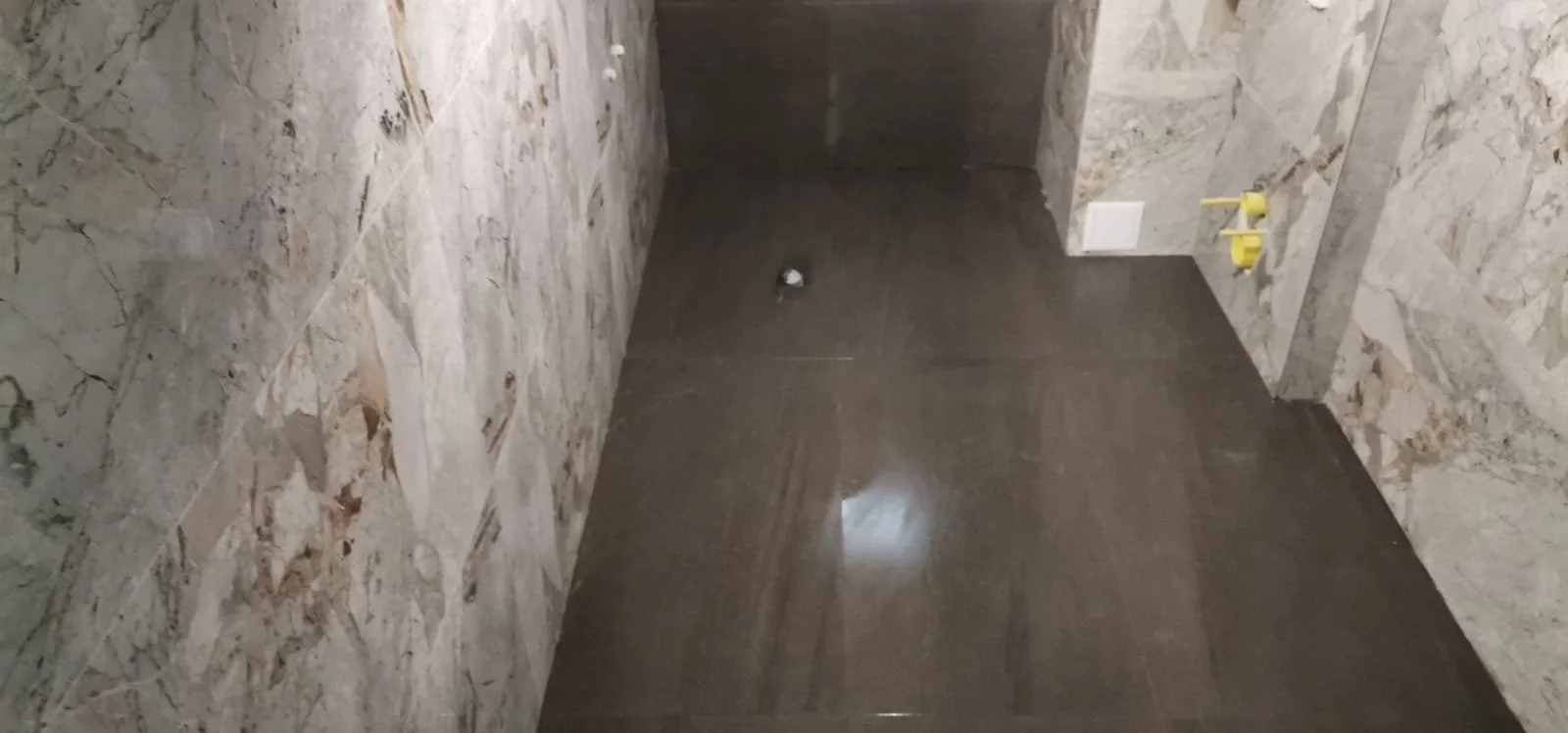 A small bathroom or laundry room with tiled walls and a dark polished floor. There is a drain in the center of the floor and yellow water supply valves on the right wall.