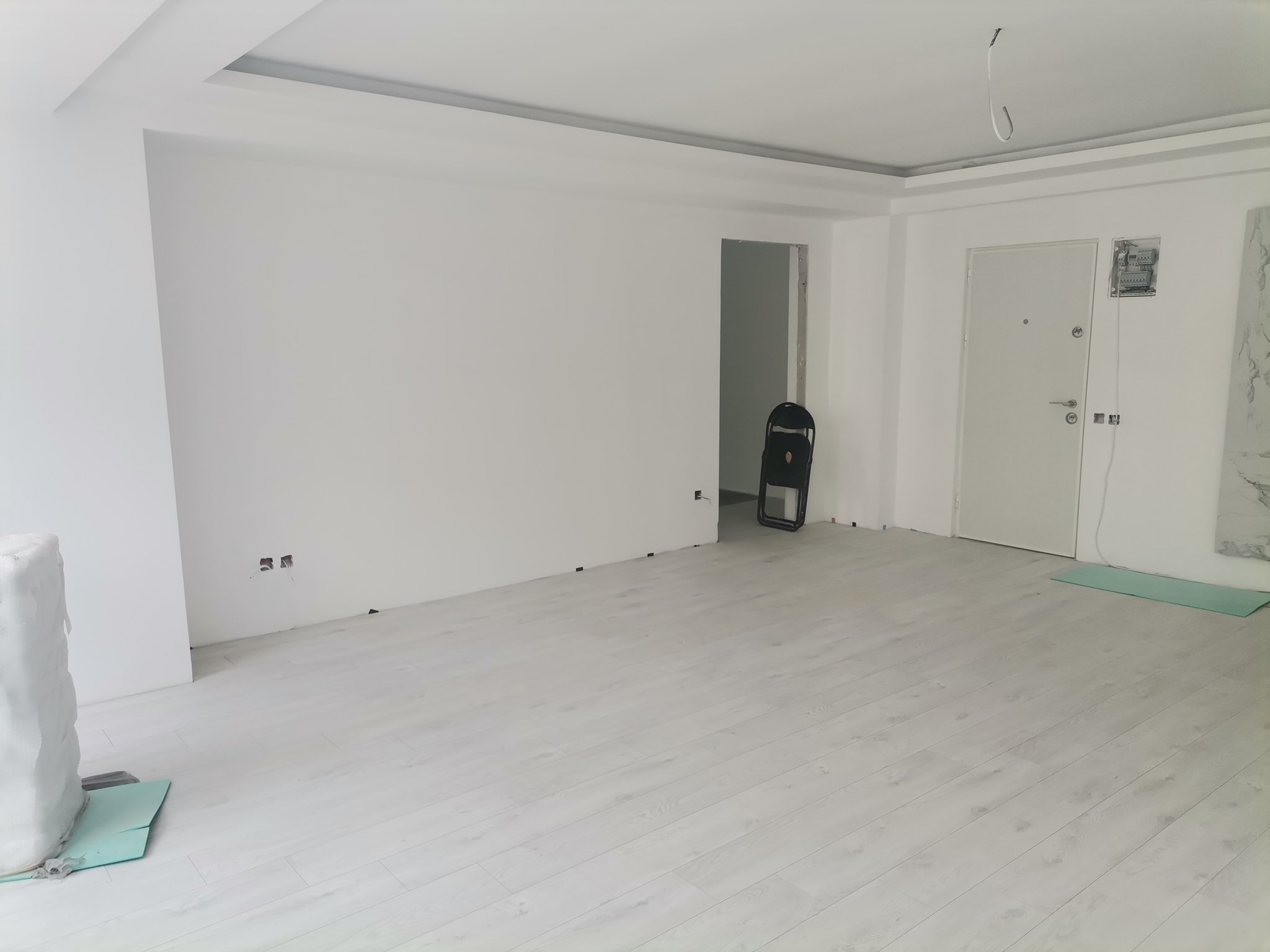 An unfinished room with white walls and a light-colored floor. There is an open doorway and an electrical panel on the wall, with exposed wiring and hooks hanging from the ceiling. There is a folding chair and some construction materials on the floor