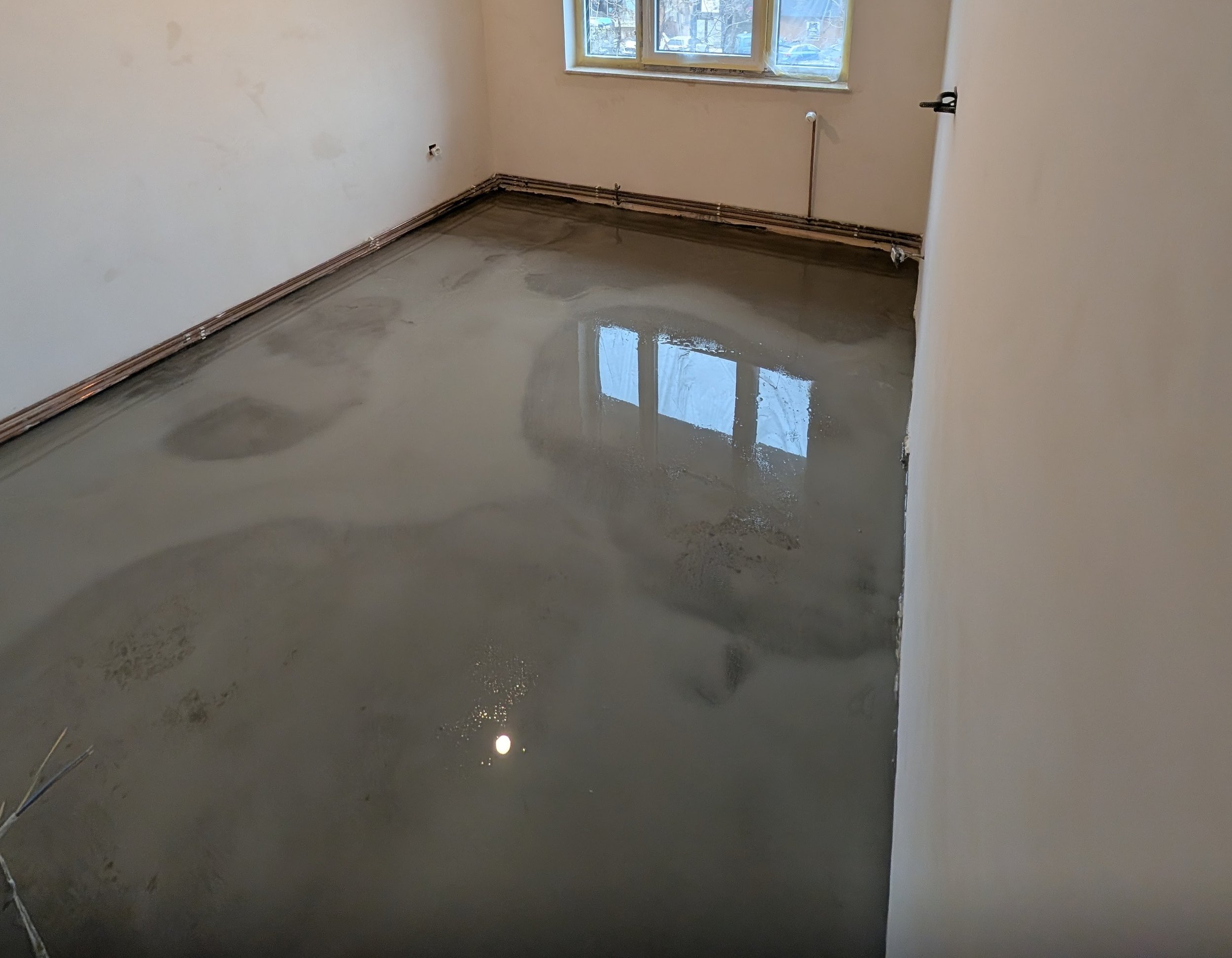 An empty room with a concrete floor that has just been poured and is still wet. The room has white walls, a window allowing natural light, and visible electrical outlets and wiring along the baseboards.