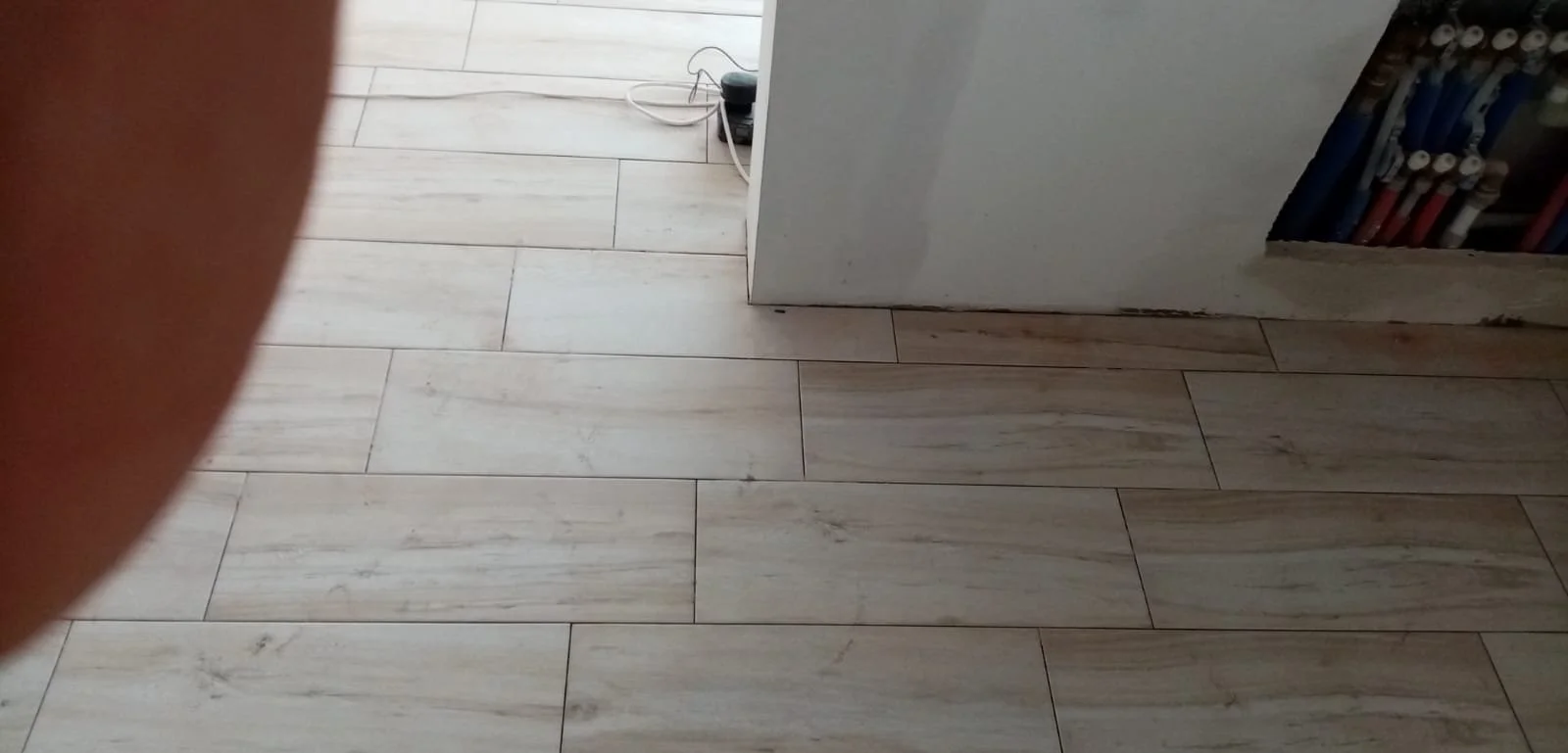 Close-up of a light-colored wooden floor, part of a white wall, with a finger partially covering the left side of the image and wires on the floor near the wall.