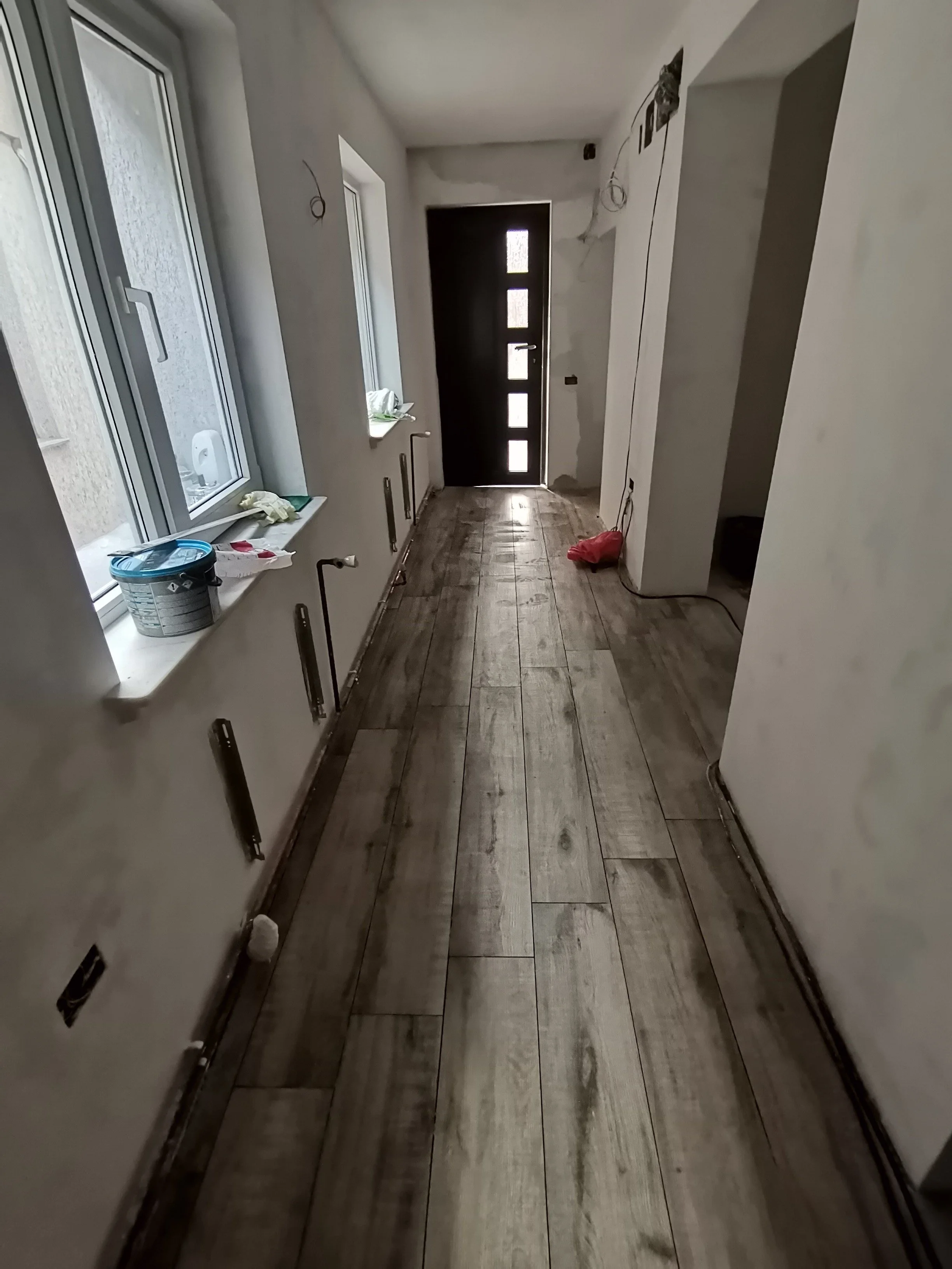 Empty interior hallway with wooden flooring, windows on the left side, and a door at the end, under construction with visible wiring and unfinished walls.
