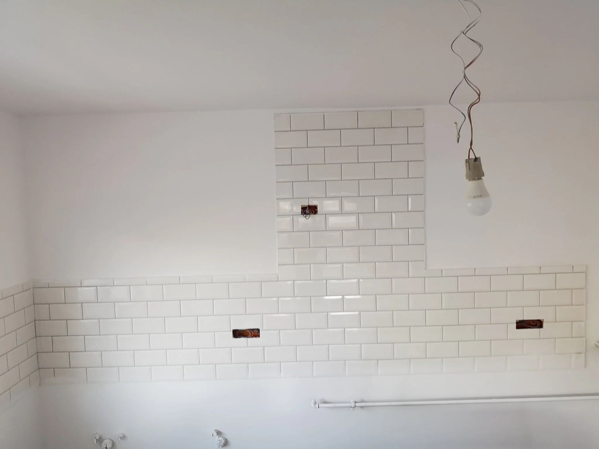 A room with white tiled walls, an unfinished ceiling with hanging light fixture wiring, and electrical outlets without cover plates.