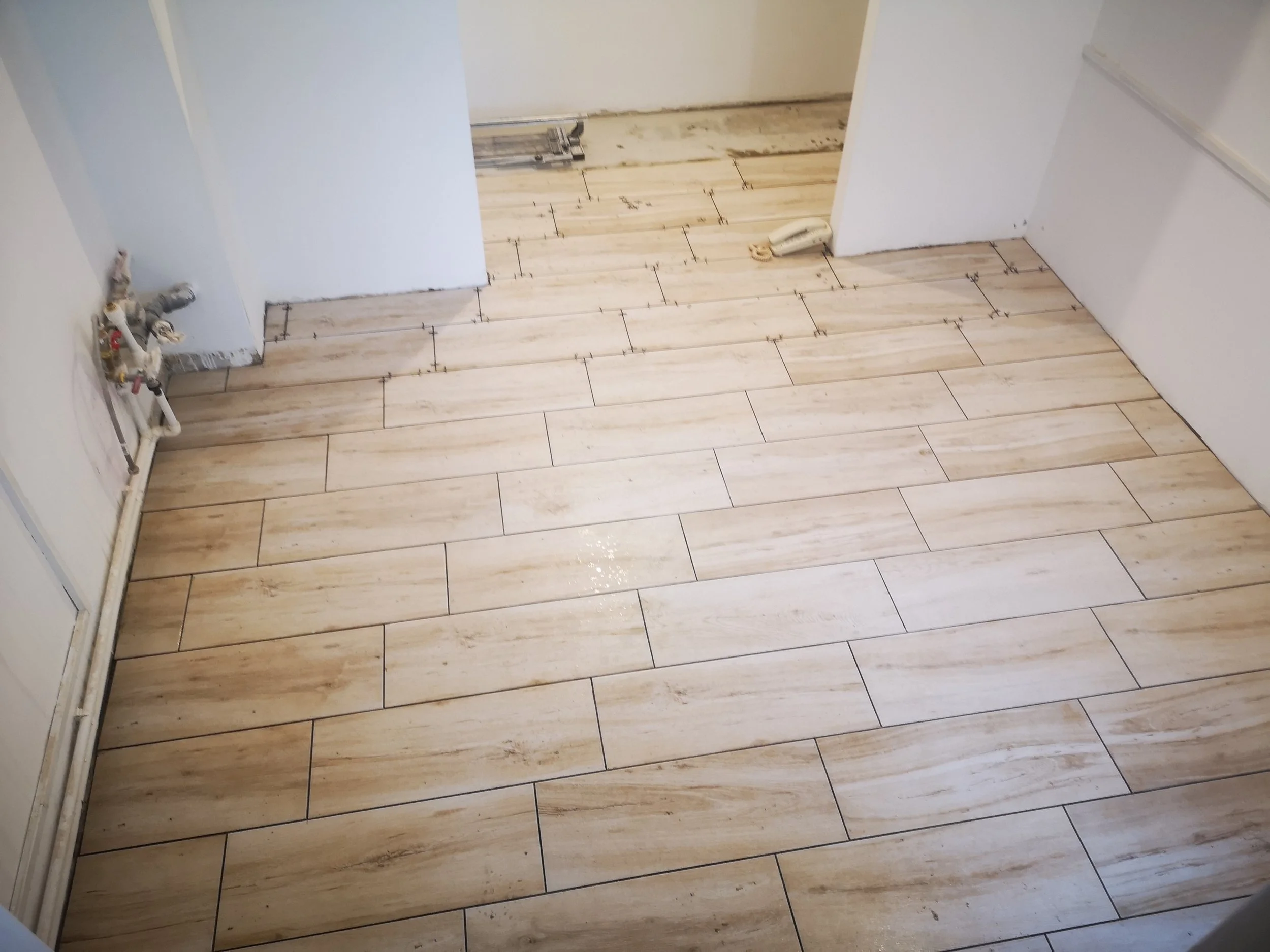 A room under construction with tiled flooring, plumbing pipes visible on the left, and an old telephone on the floor near the doorframe.