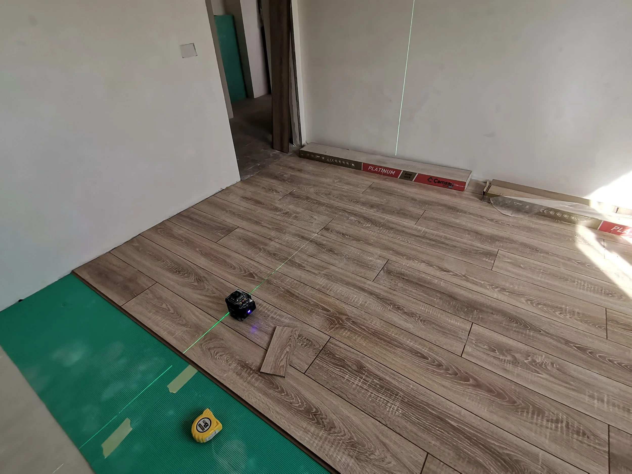 Interior of a room under construction with new wood flooring, a green laser level string on the floor, a yellow measuring tape, and construction materials including boxes and panels along the walls.