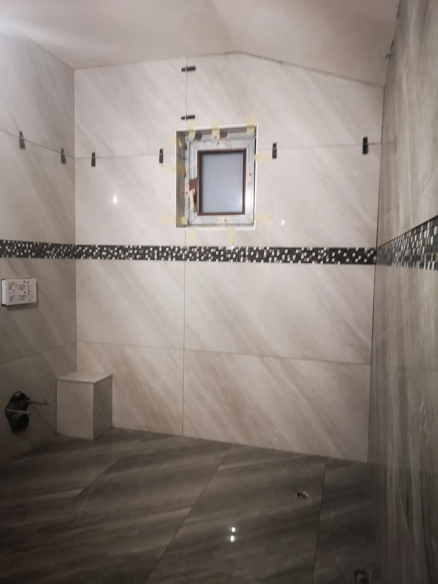 Interior view of a modern bathroom under construction, with large light-colored tiled walls, a small frosted window, black clamps holding tiles, and a strip of mosaic tiles running vertically.