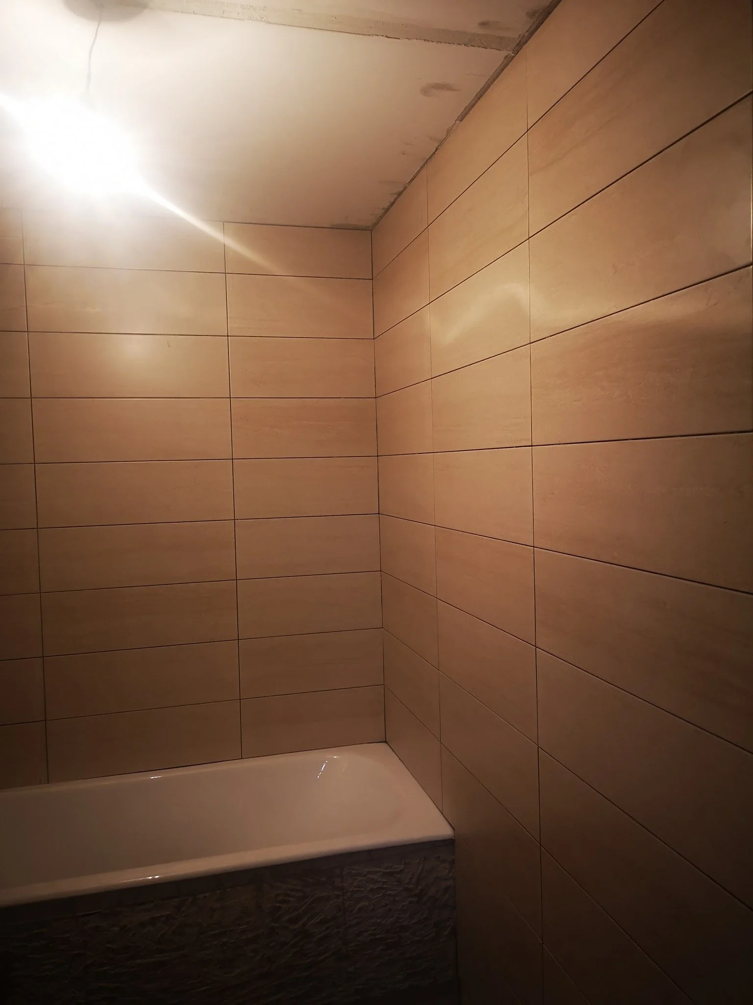 A corner of a bathroom with beige ceramic tiles on the wall and a white bathtub at the bottom. The ceiling appears unfinished or under construction.