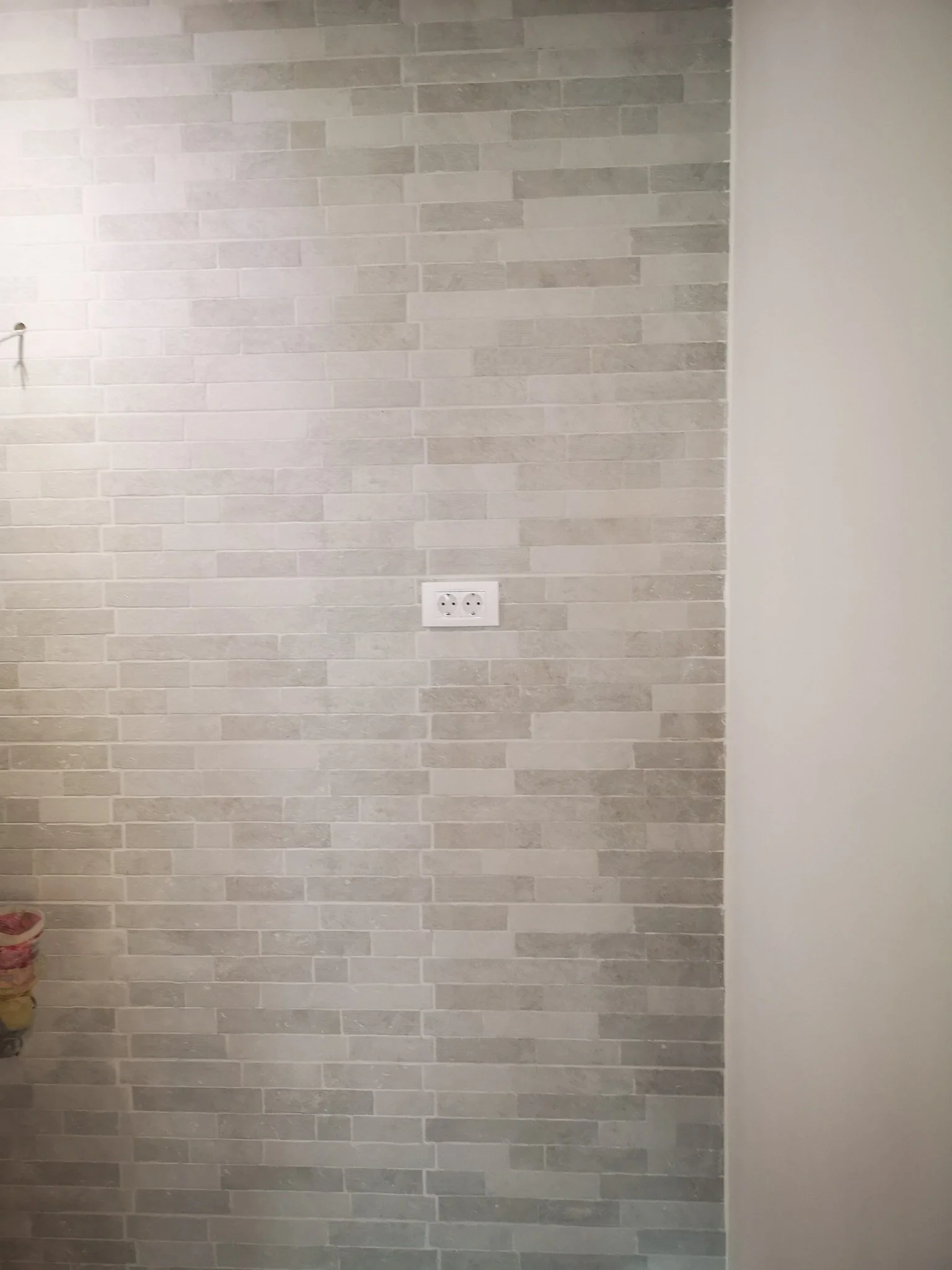 A wall with light gray brick tiles and a white wall on the right side, with an electrical outlet in the middle.