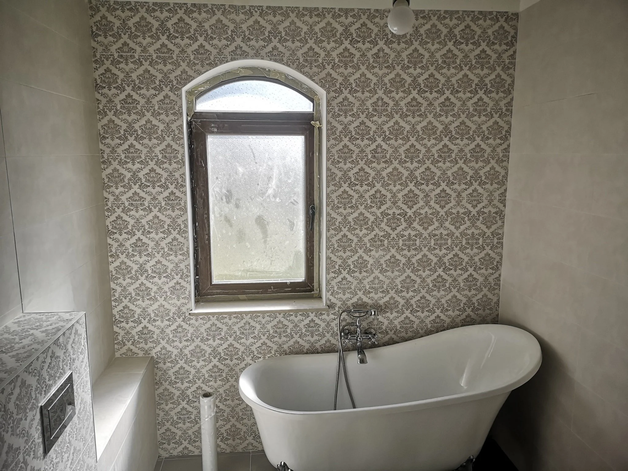 A bathroom with gray damask-patterned tiles on the wall, a white bathtub, a silver faucet, a window with frosted glass, and beige tiles on the adjacent wall.