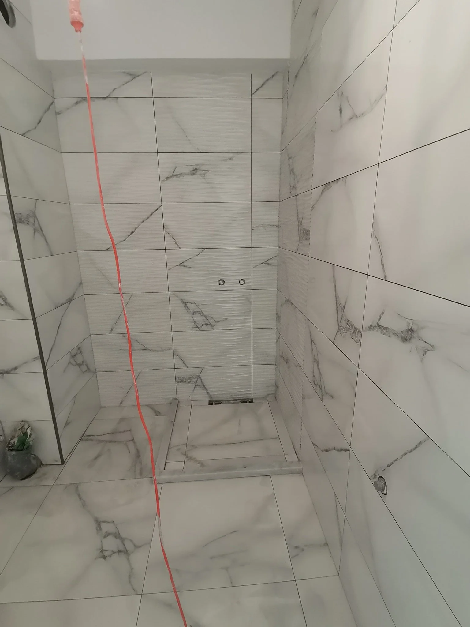 A bathroom with white marble tiles on the walls and floor. There is a drain on the floor, and a red string or line running vertically across the image. Some construction or renovation materials are visible in the corner.