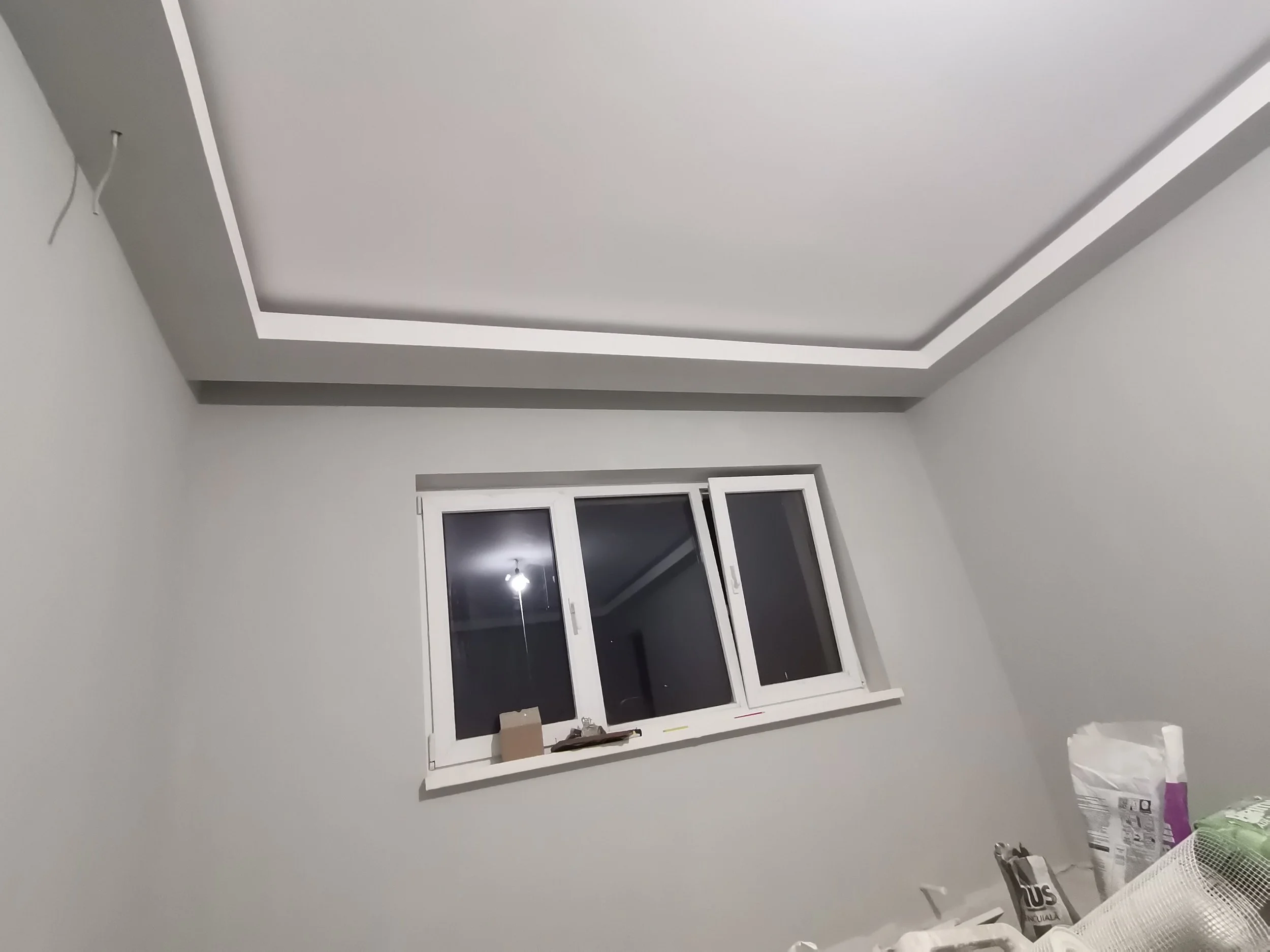 An unfinished room with a ceiling featuring a recessed tray design. There is a window with a white frame on one wall, and some items including boxes and bags are on a table or surface in the foreground.