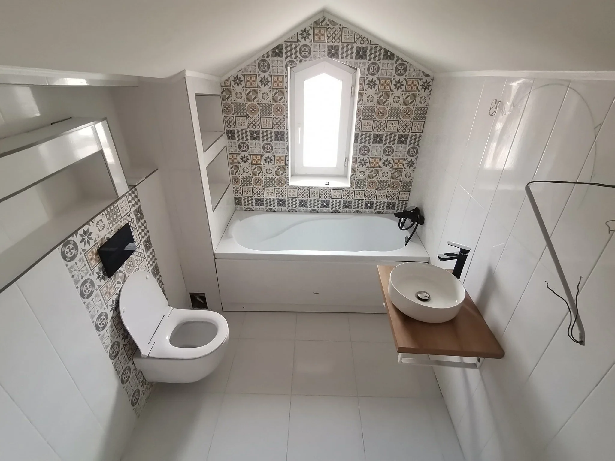 A bathroom with white tiled walls and floor, a white toilet, and a white bathtub. There are open shelves on the left wall and a window with patterned tiles around it on the back wall. A black hairdryer is hanging in the corner, and a wooden table wit