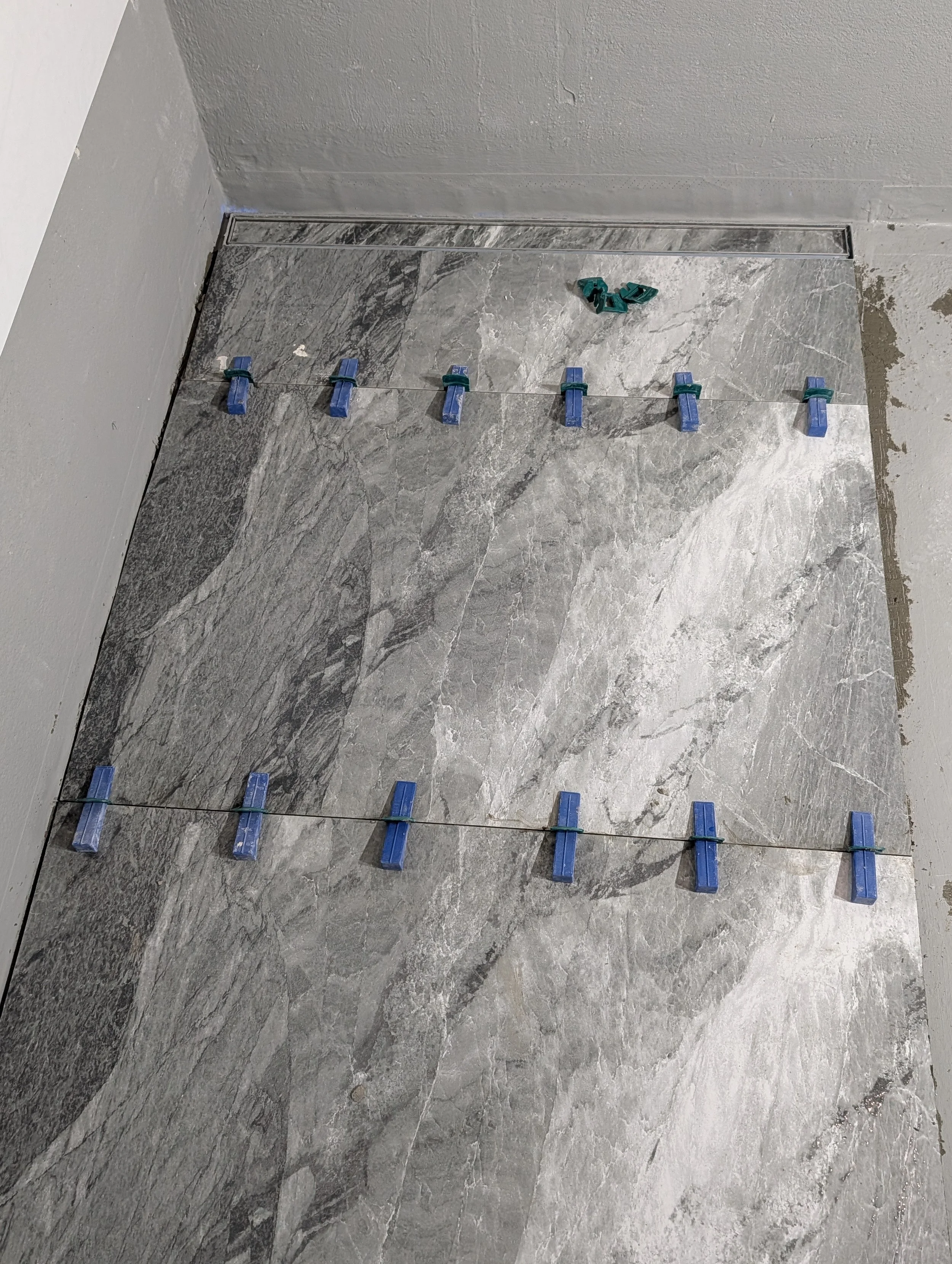 Gray marble floor with rows of blue and green tile spacers and cross spacers used for tiling, with a trowel and other small tools on the floor.
