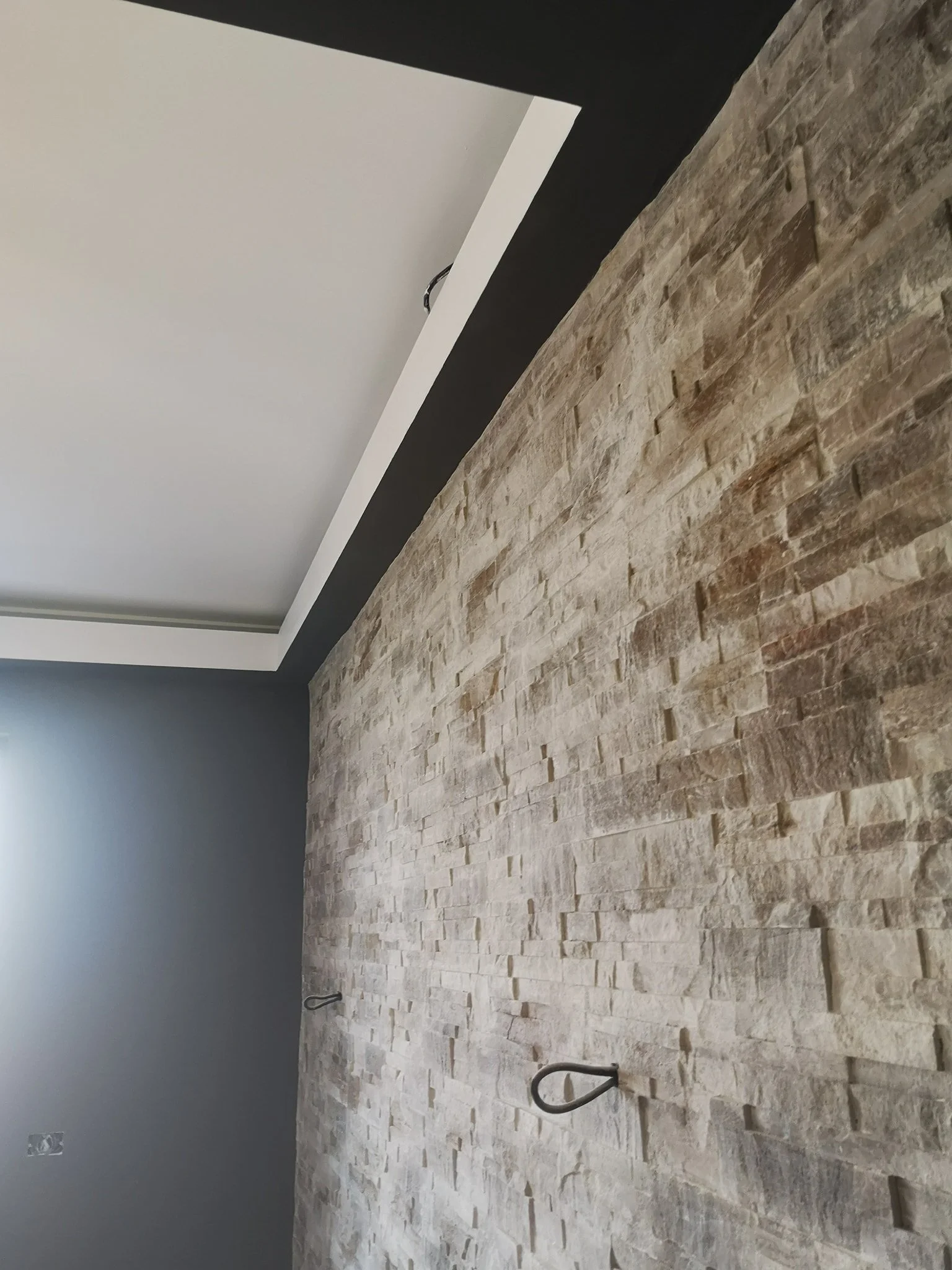 A room under construction or renovation with a beige brick wall, a gray painted wall, and a sloped ceiling with a black border. There are black metal hooks or handles attached to the brick wall, and an electrical outlet is visible near the floor.