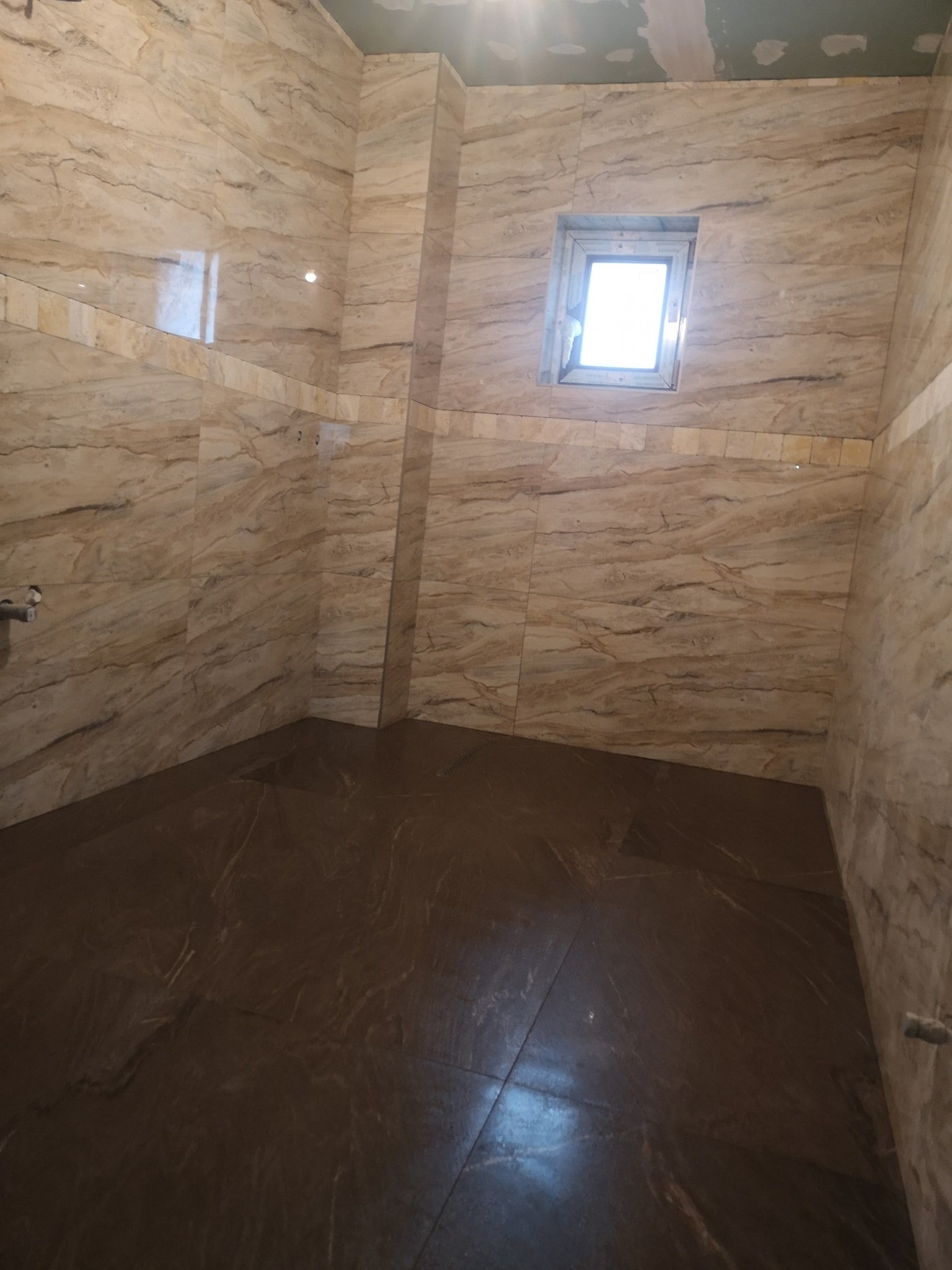 Interior of a bathroom shower with beige marble tiles on the walls and dark brown tiles on the floor. There is a small window on the wall.