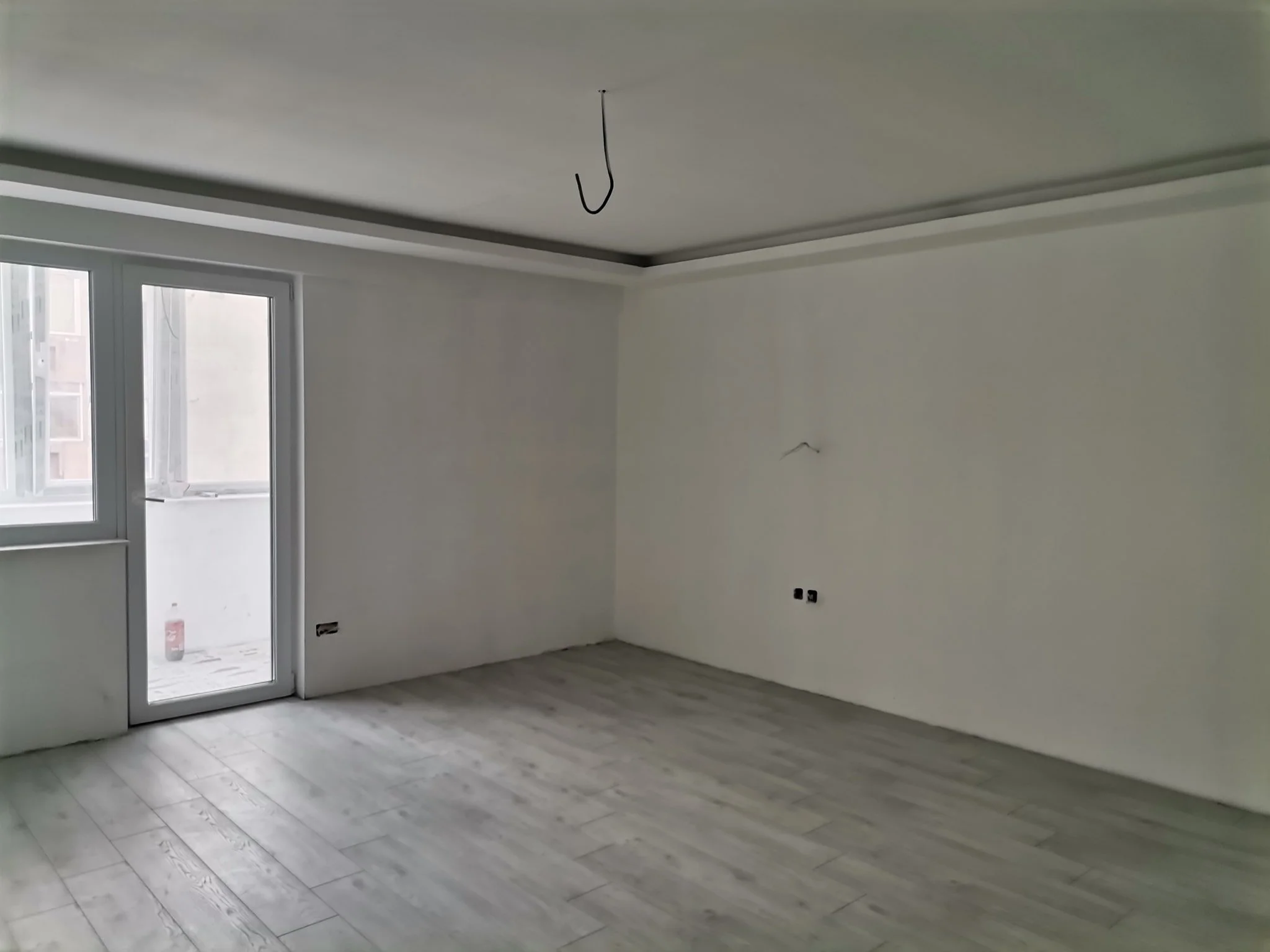 Empty room with light wood flooring, white walls, large window, and door leading to balcony, with a hanging wire from ceiling.
