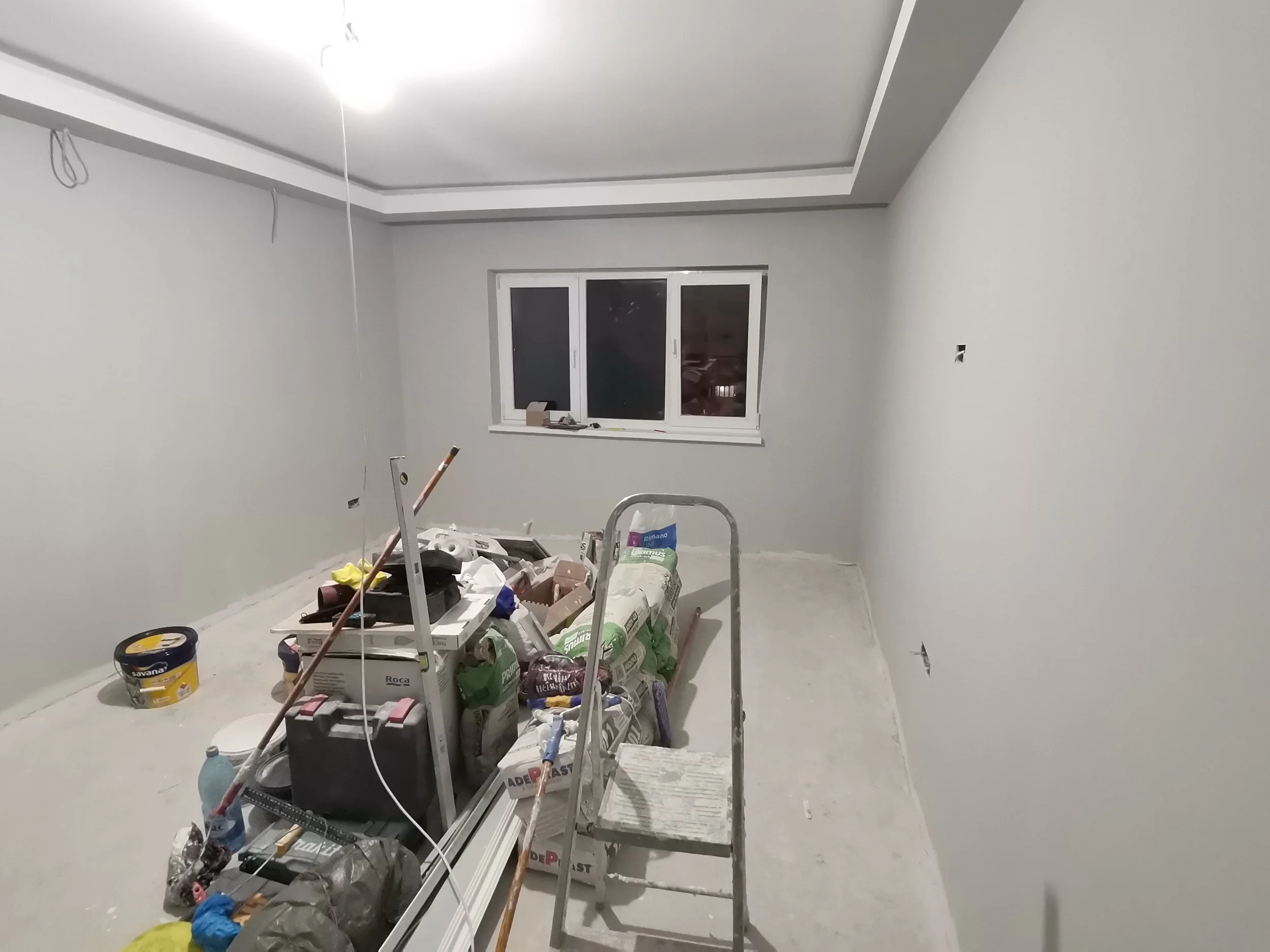 Empty room under renovation with construction supplies, drywall, tools, and a window.