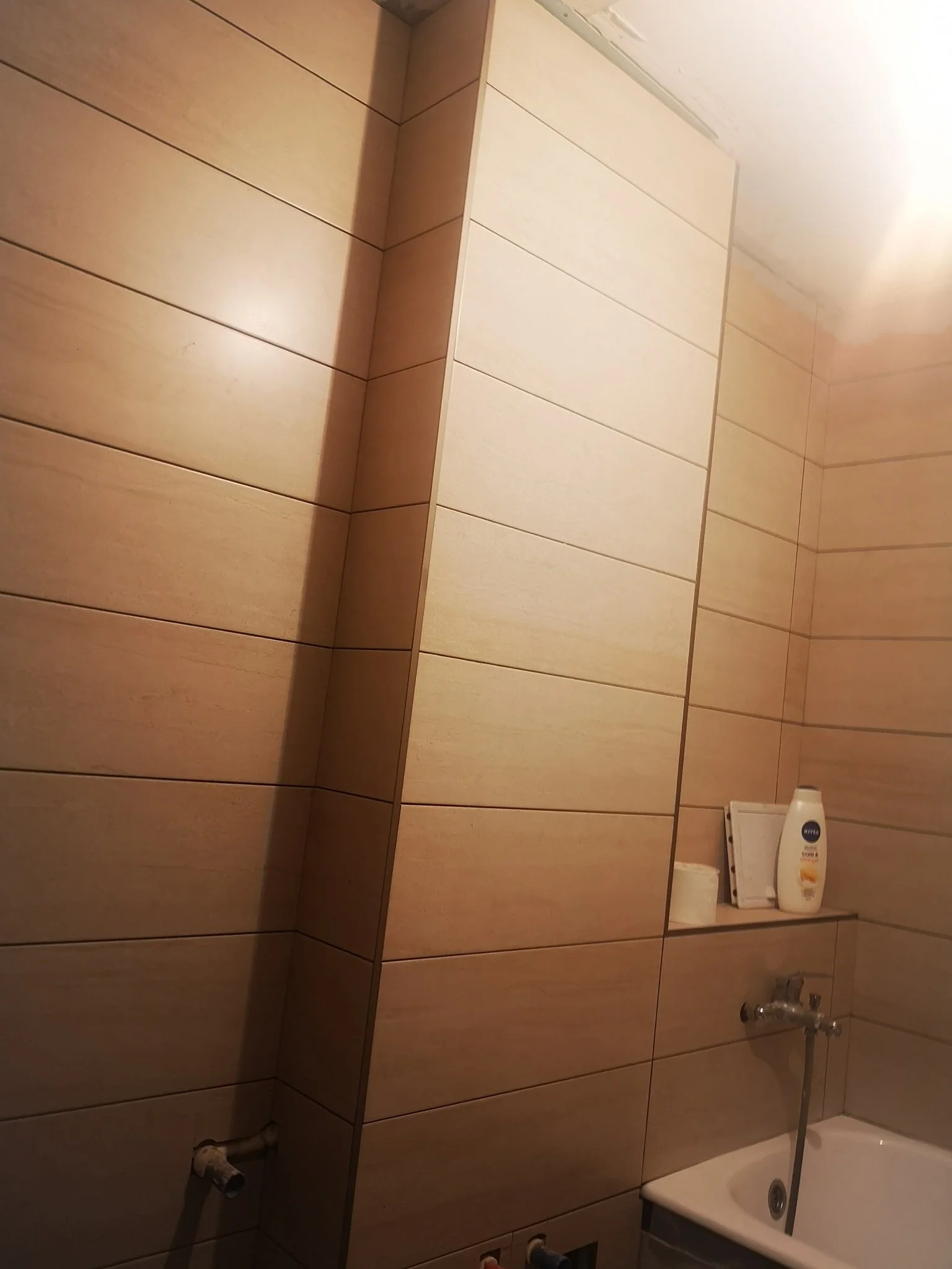 Bathroom with beige tiled walls, a white bathtub, a bottle of lotion, and a toilet paper roll on a shelf.