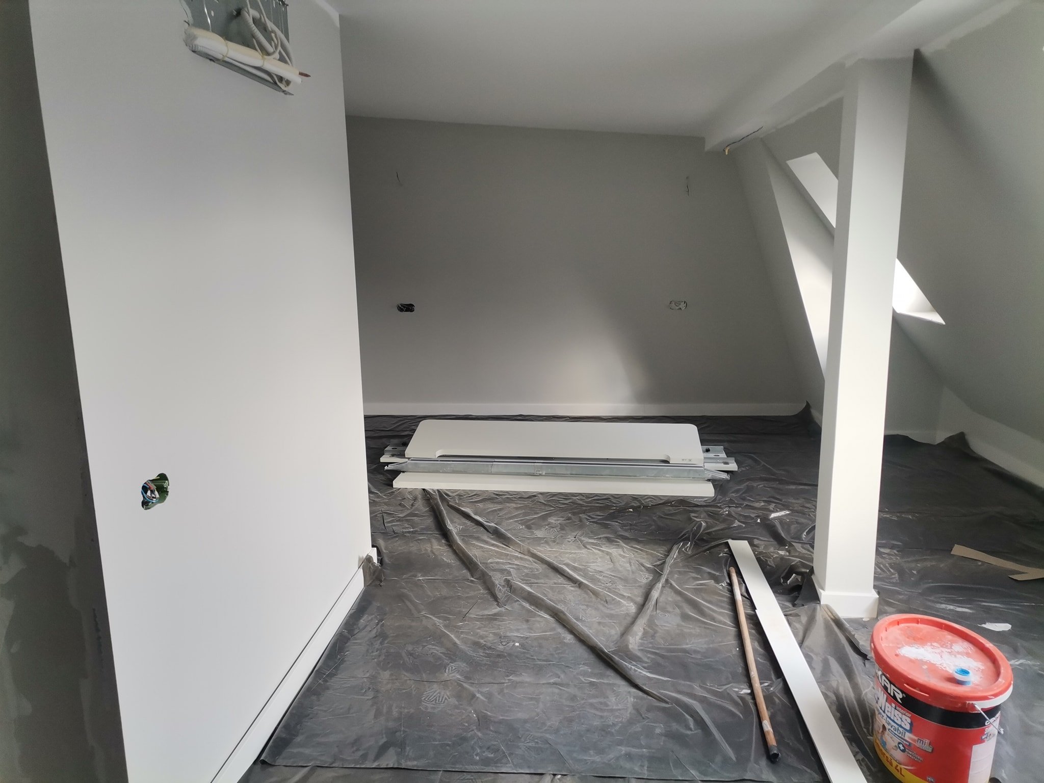 An unfinished room under construction or renovation. There are various construction tools and materials on the floor, including a paint bucket, a putty knife, and some metal framing. The walls are painted gray, and there are electrical outlets and wi
