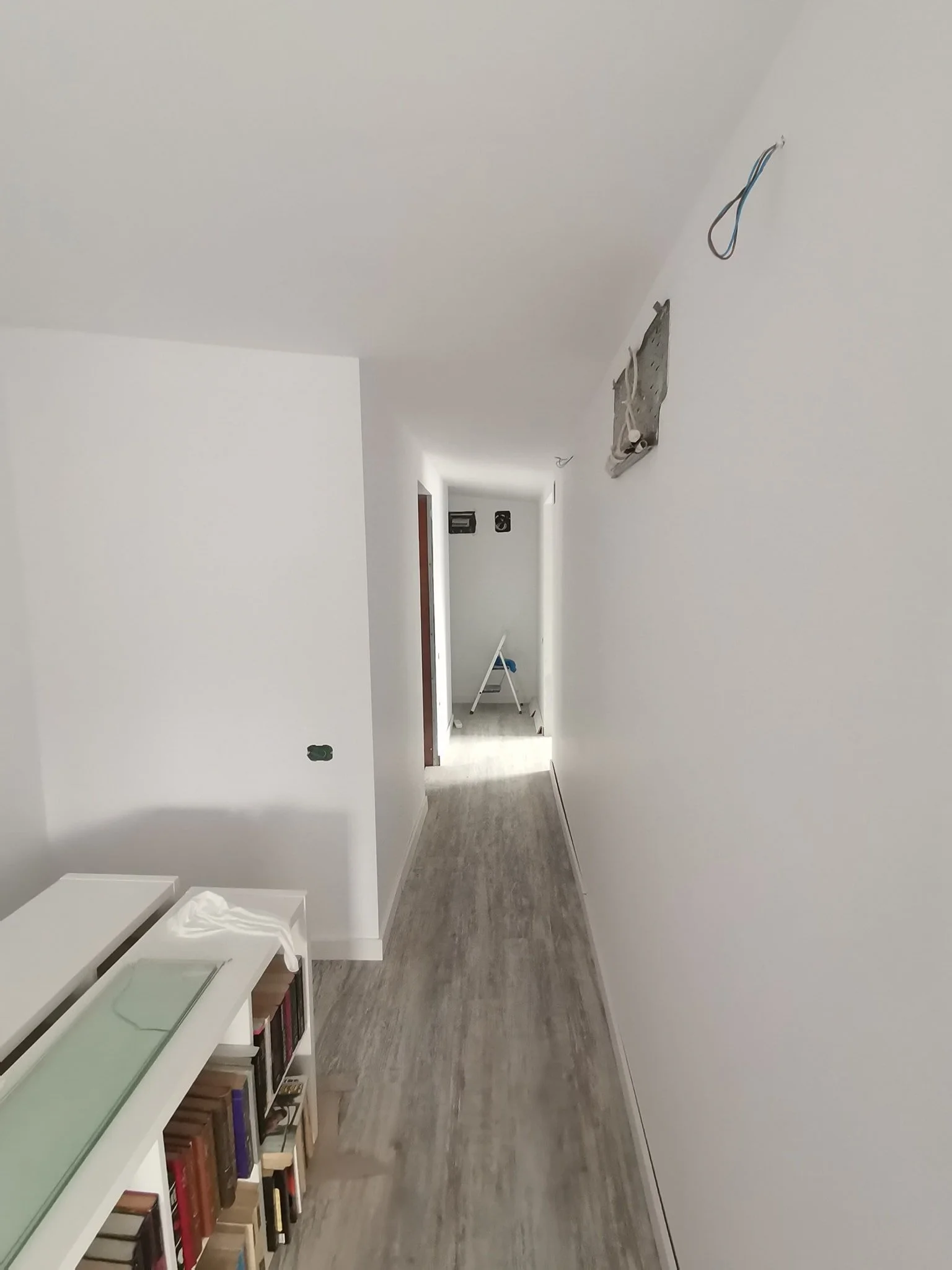 A hallway under construction with white walls and a wood floor, featuring an open bookshelf on the left side and a partially installed light fixture on the ceiling.