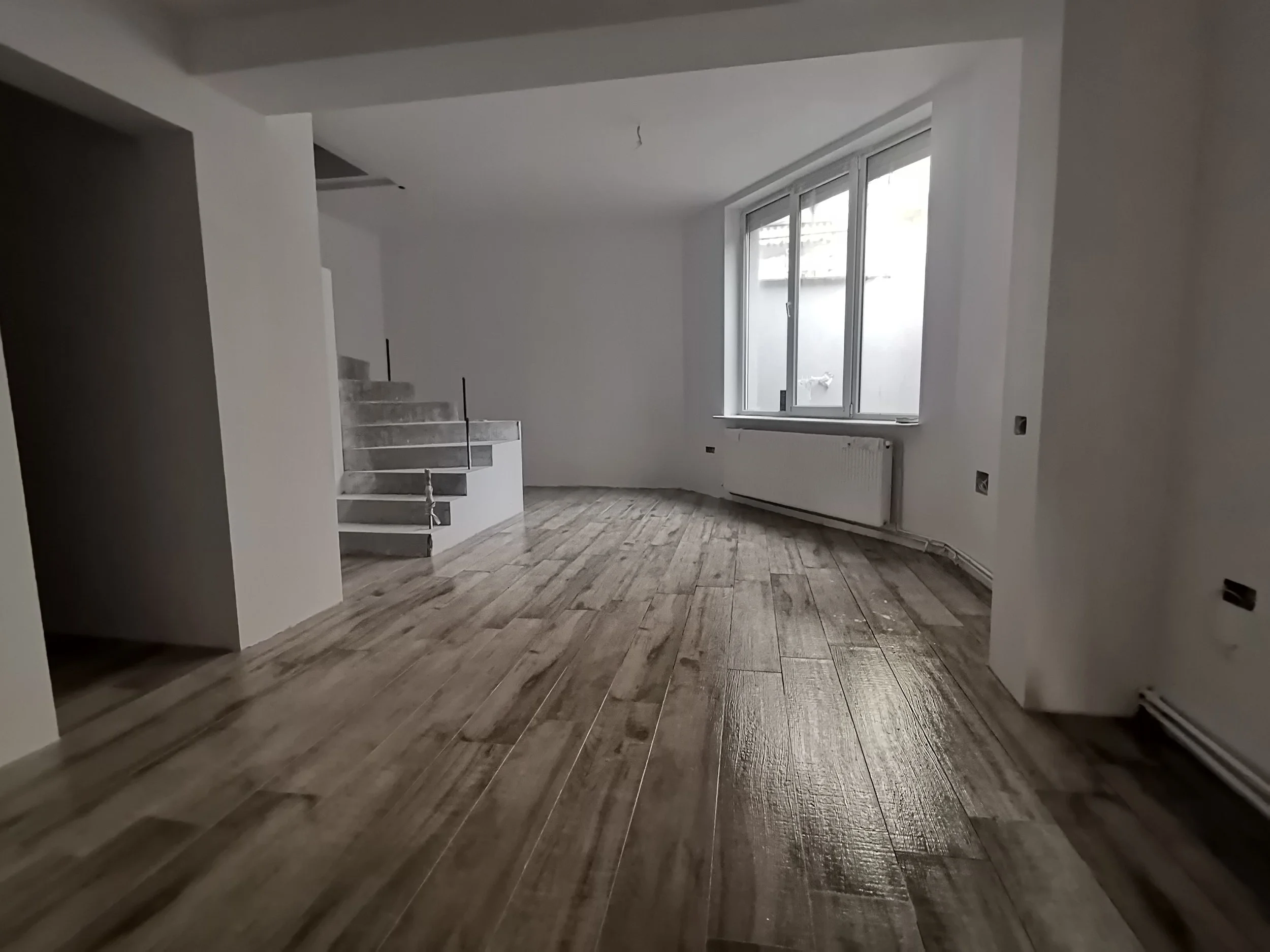 Empty room with hardwood flooring, white walls, a large window, and a staircase.