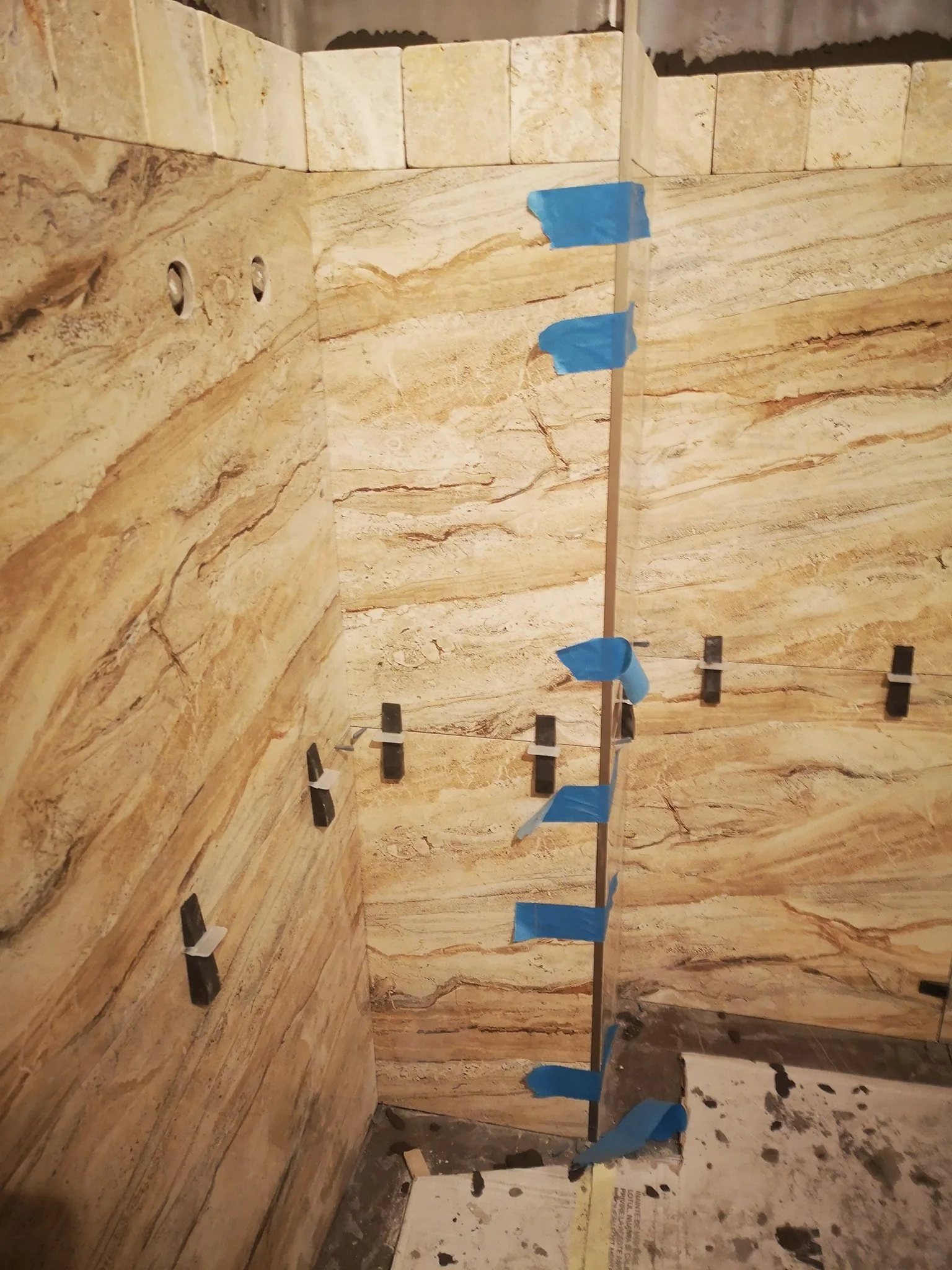 Interior of a room with walls covered in beige and brown marble tiles. A vertical strip of blue painter's tape is attached to a metal rod running from top to bottom. Several black and white plastic spacers are attached at the bottom of the rod. The f