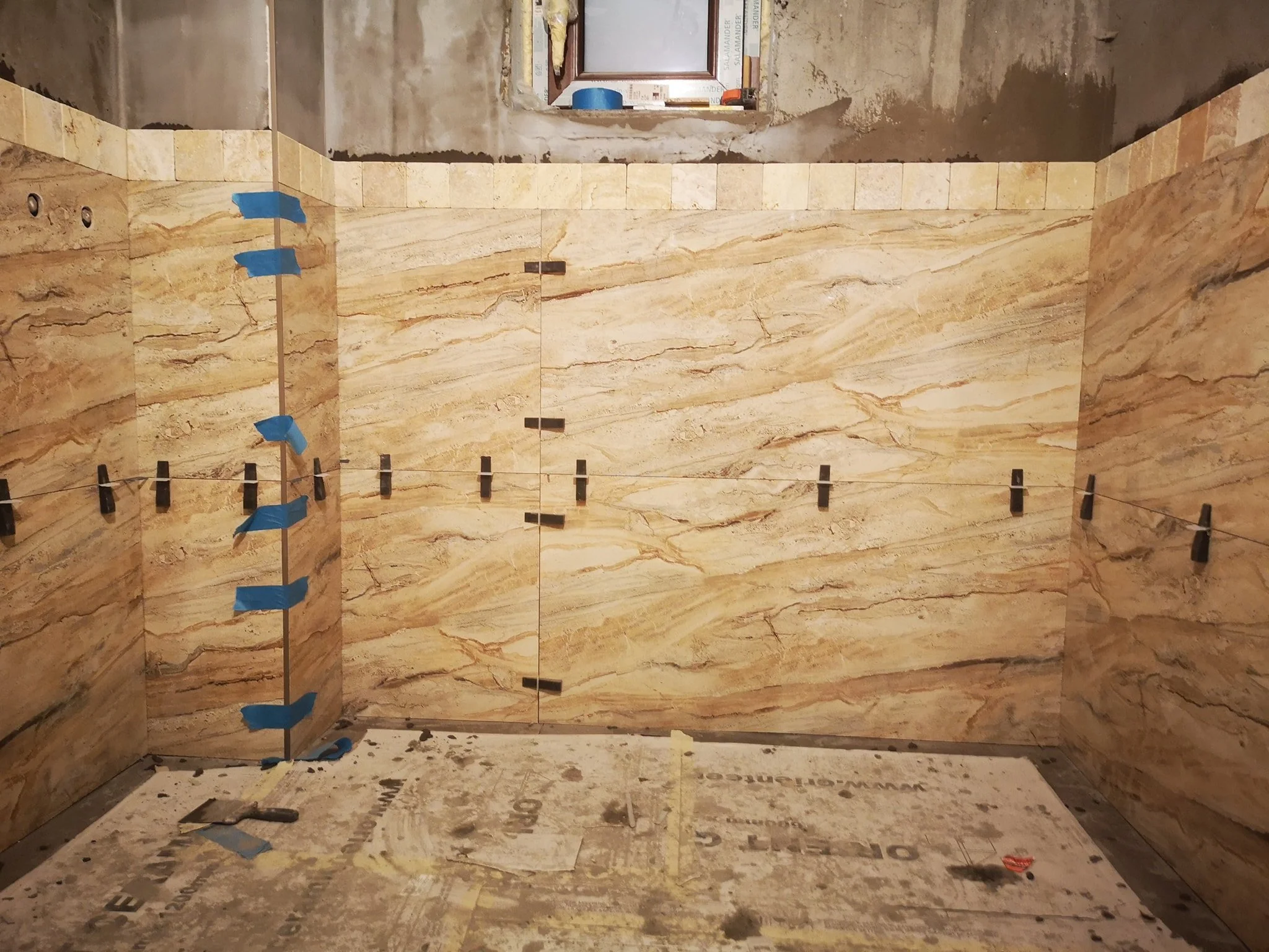 Bathroom under construction with large beige marble tiles on the walls, partially tiled shower area, waterproofing panels, and construction tools and materials scattered on the floor.