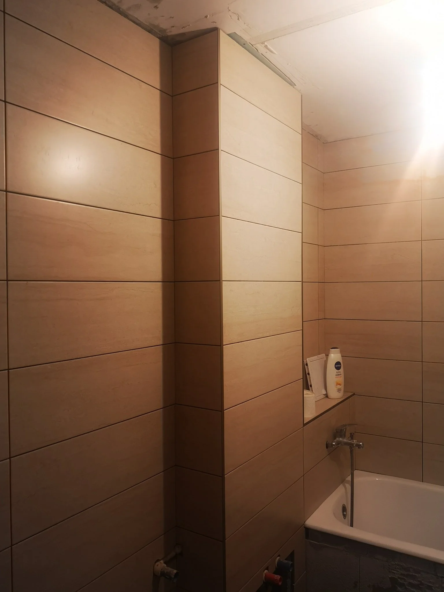 Bathroom with beige tiled walls, a small white bathtub, a shelf holding shampoo and soap, and visible plumbing fixtures.
