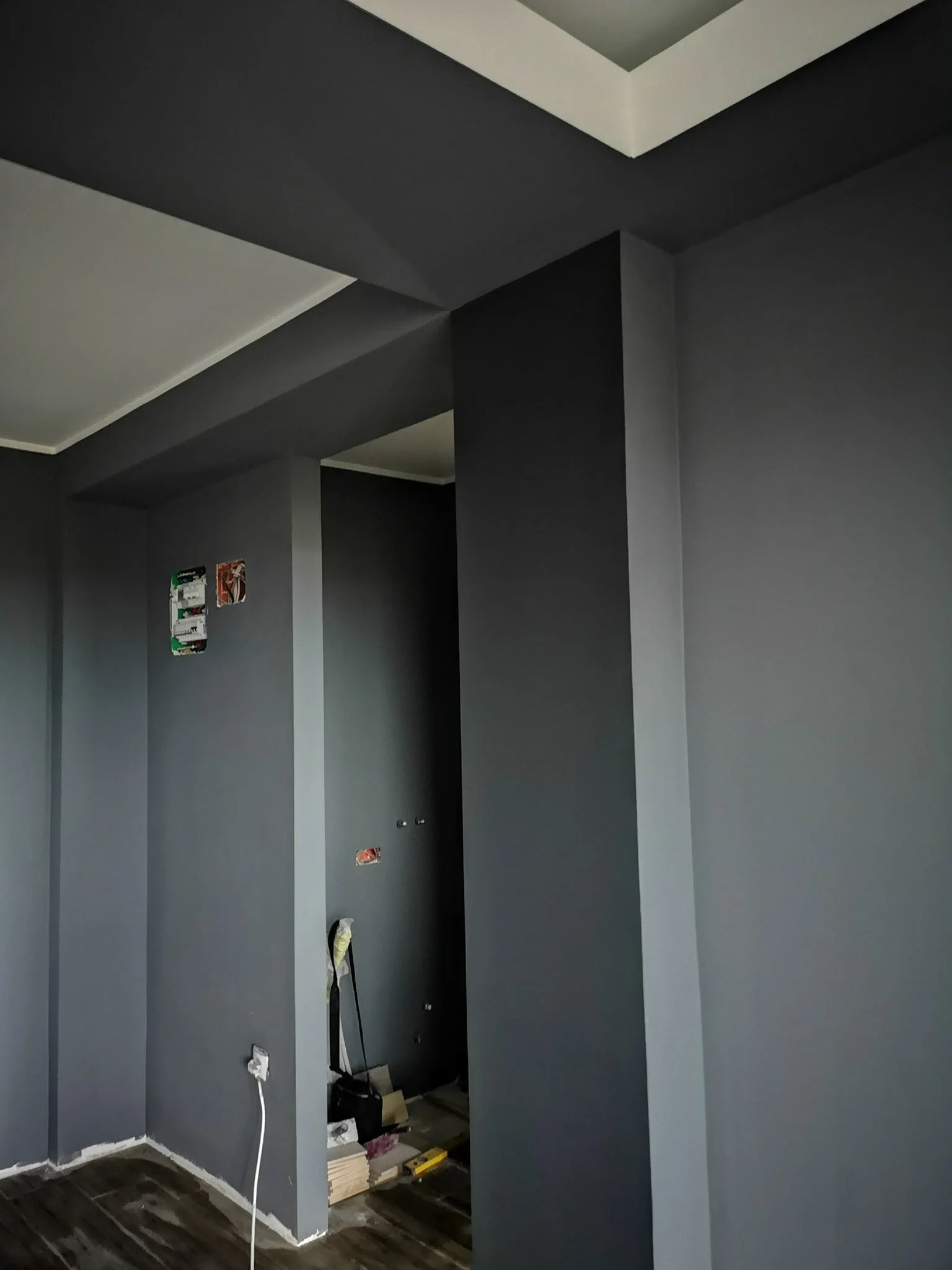 Interior of a room under construction, with gray walls and a ceiling, electrical outlets, and wires hanging. Construction materials are scattered on the floor.