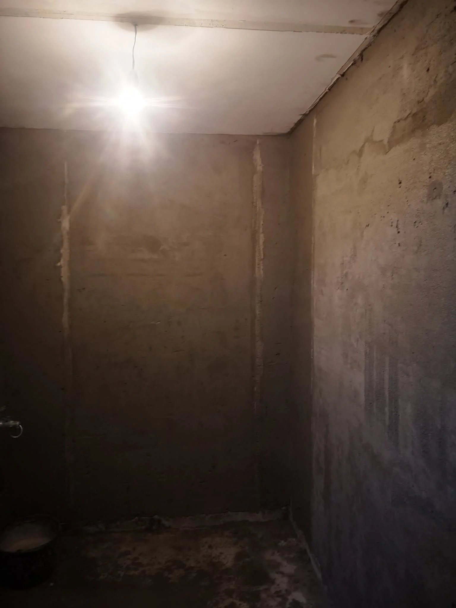 Empty room with unfinished walls and floor, a single bright ceiling light, and visible electrical wiring.