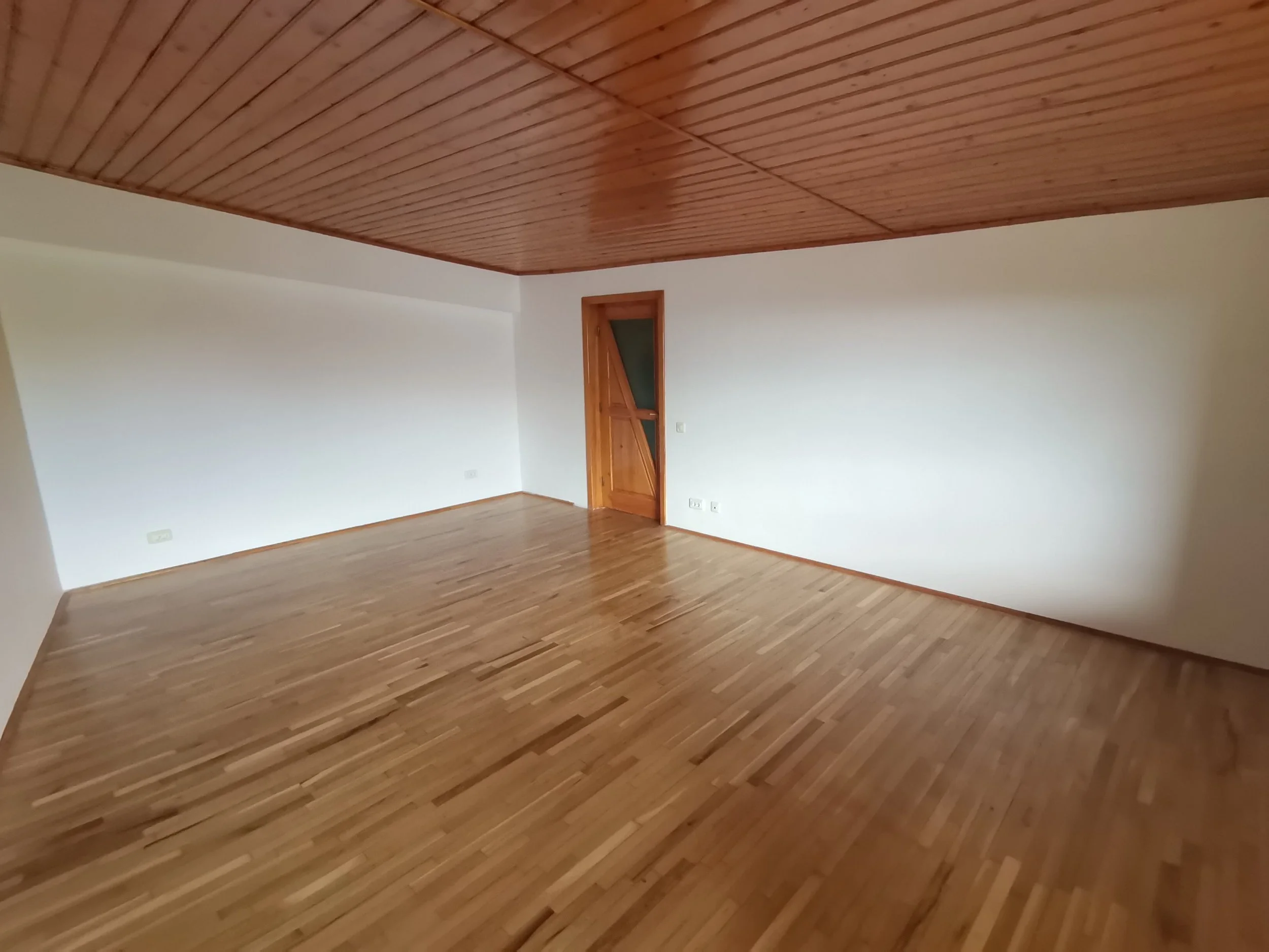 An empty room with a wooden ceiling, white walls, and a parquet floor. There is a closed wooden door on one wall.