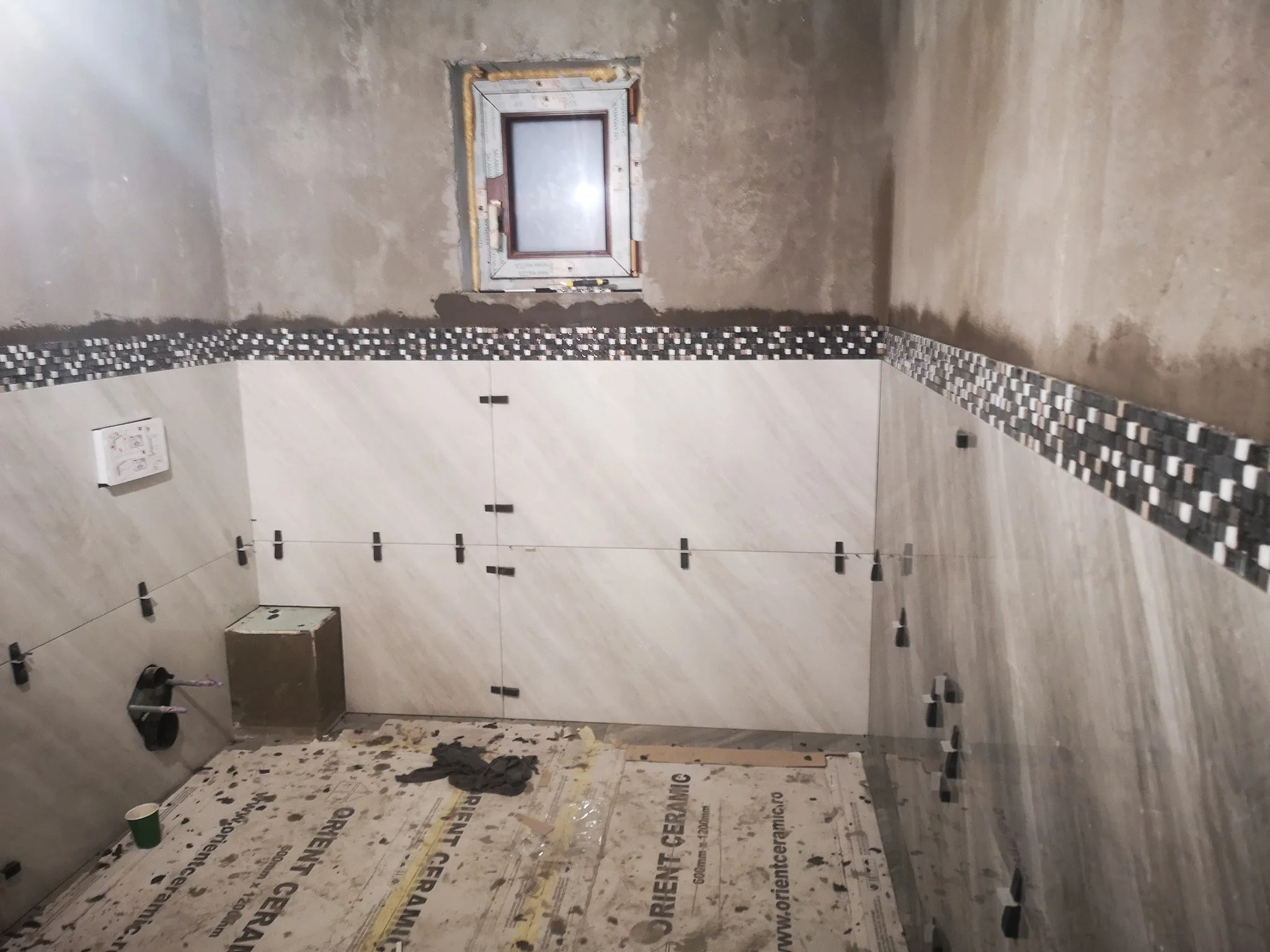 Unfinished bathroom with tiled walls and floor. The wall tiles are partially installed, with some tiles missing and exposed drywall and insulation. There is a small window in the wall. The floor has protective covering and scattered construction debr
