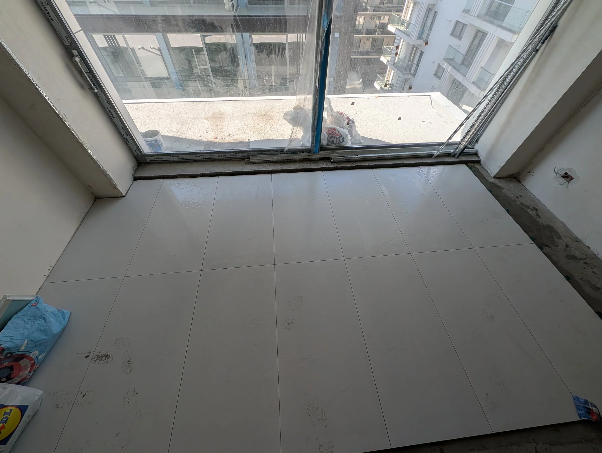 View from inside an apartment under construction, looking out through a glass door to a balcony, with construction materials inside and neighboring apartment buildings outside.